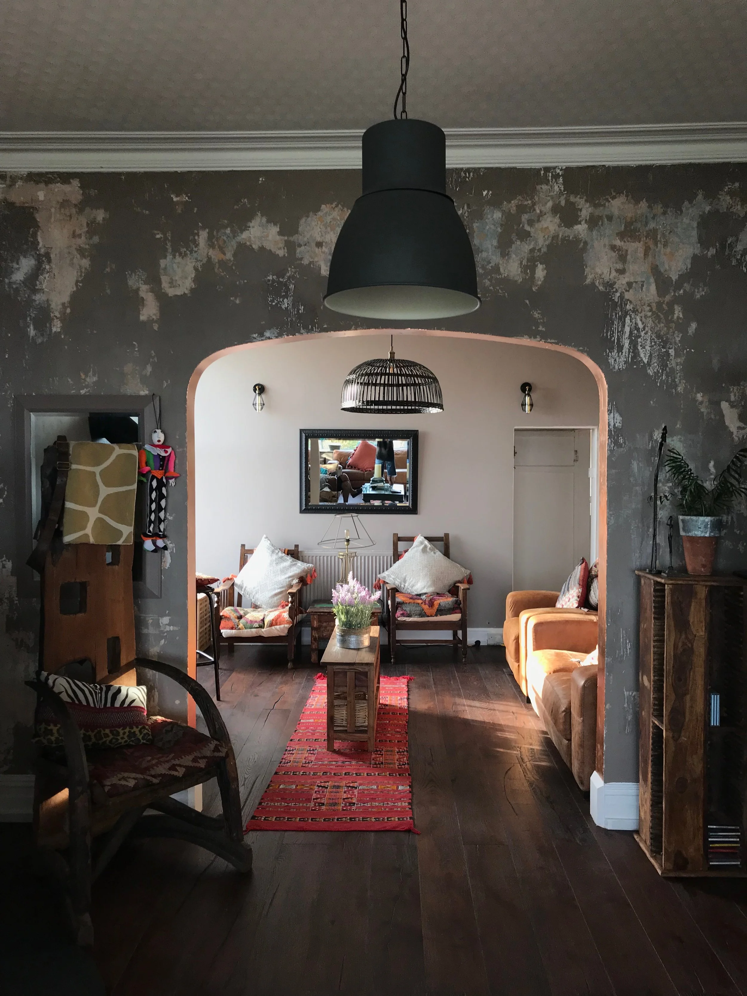 Interior living room with a dark wooden floor, a central red patterned rug, and various chairs and a sofa. The room features a large mirror on the back wall, a small table with a flower arrangement, and wall-mounted lights. The entryway has an arch with a light-colored wall and textured gray wall on the sides, and there is a black ceiling light fixture.