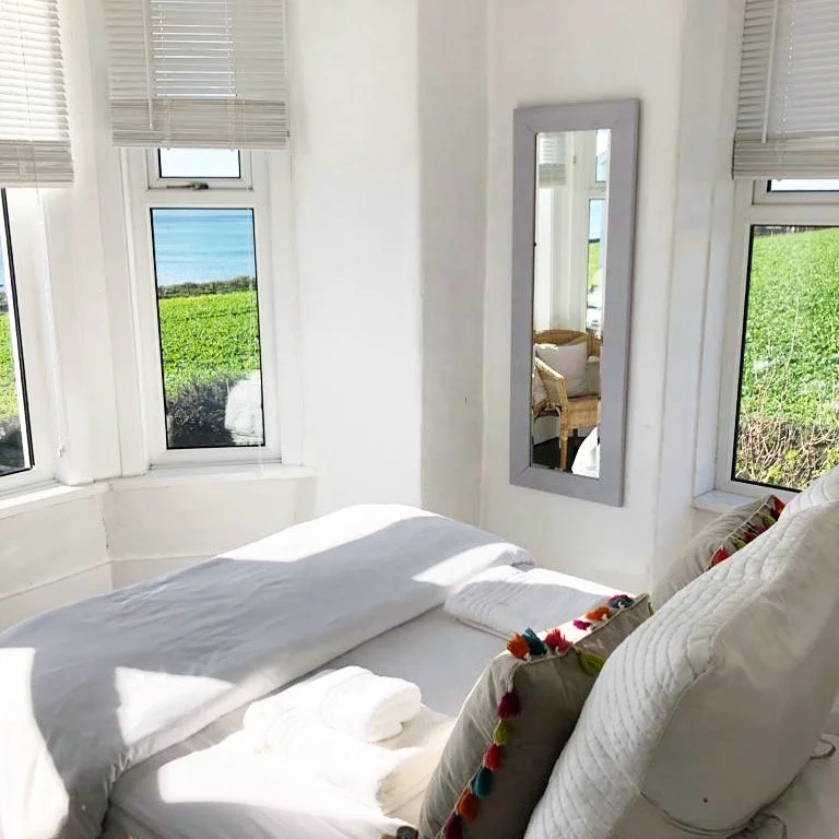 Cozy bedroom with white bedding, a mirror on the wall, and windows showing a view of the ocean and green plants outside.