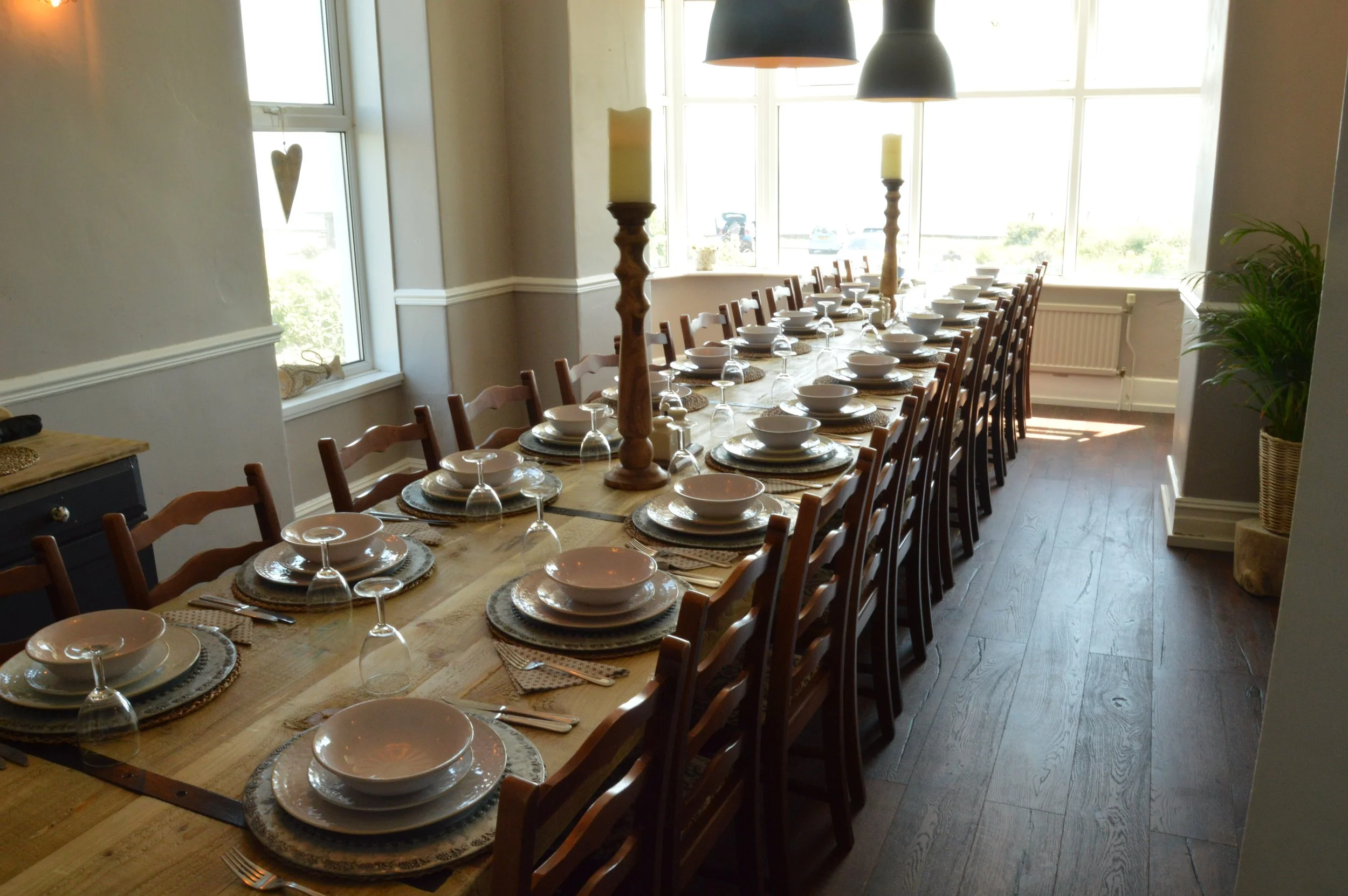 A long dining table set for a meal with bowls, plates, glasses, and utensils, with two tall candleholders in the center, in a bright room with large windows.