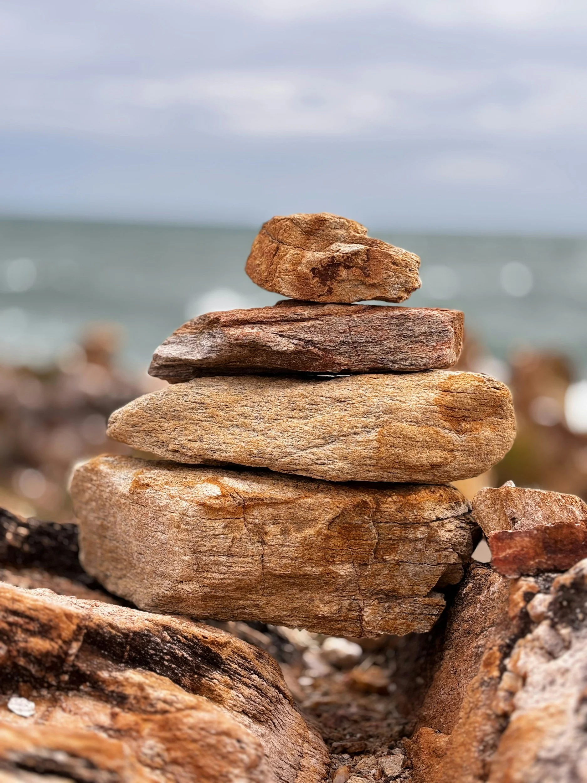Close-up of a stack of five flat, uneven rocks or stones arranged on top of each other near a body of water, with a cloudy sky in the background.