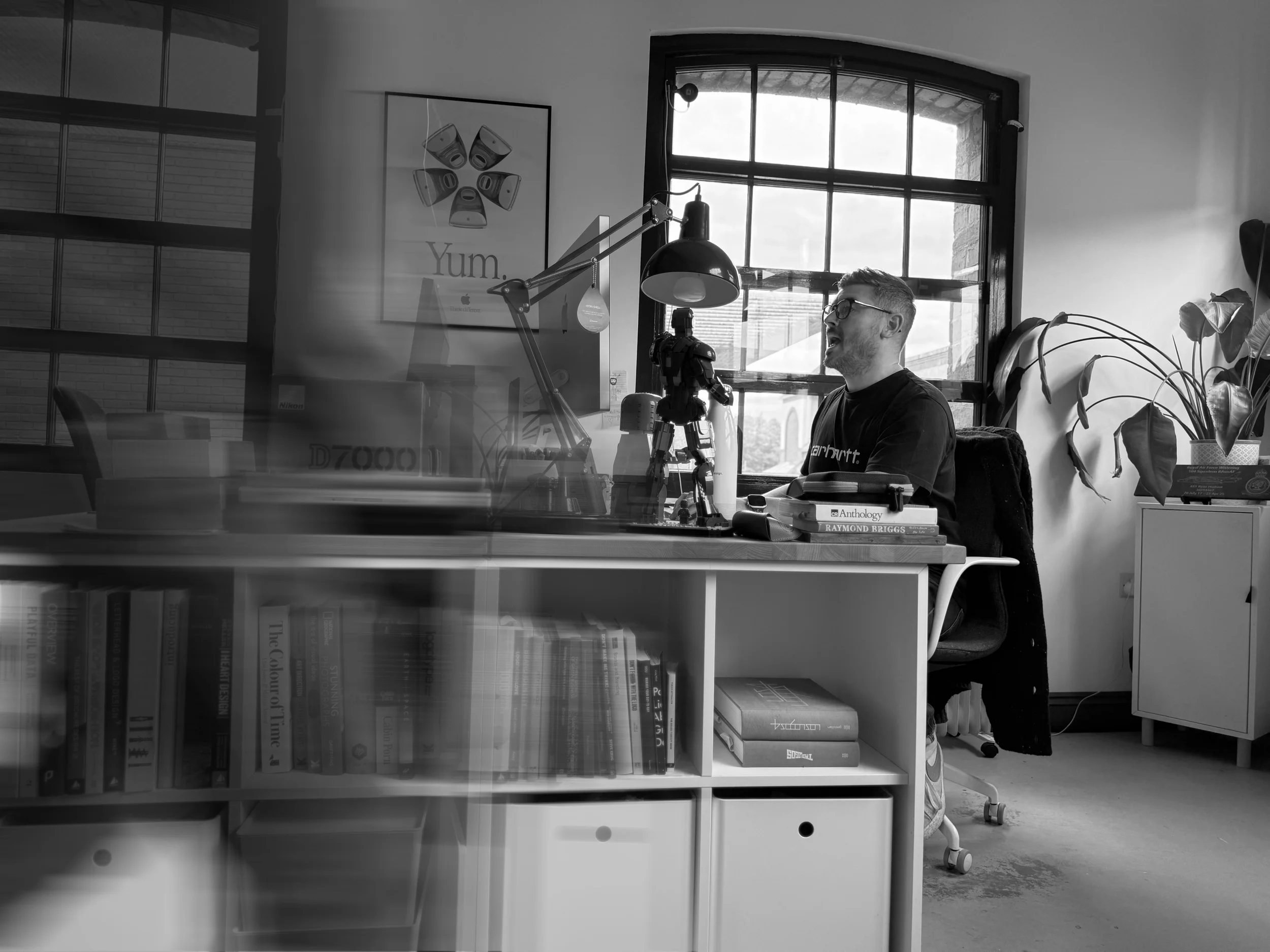 A man with glasses and a beard sitting at a desk in a modern office, speaking or singing into a small robot figure, with a window showing a cloudy sky behind him, a plant, and shelves filled with books.