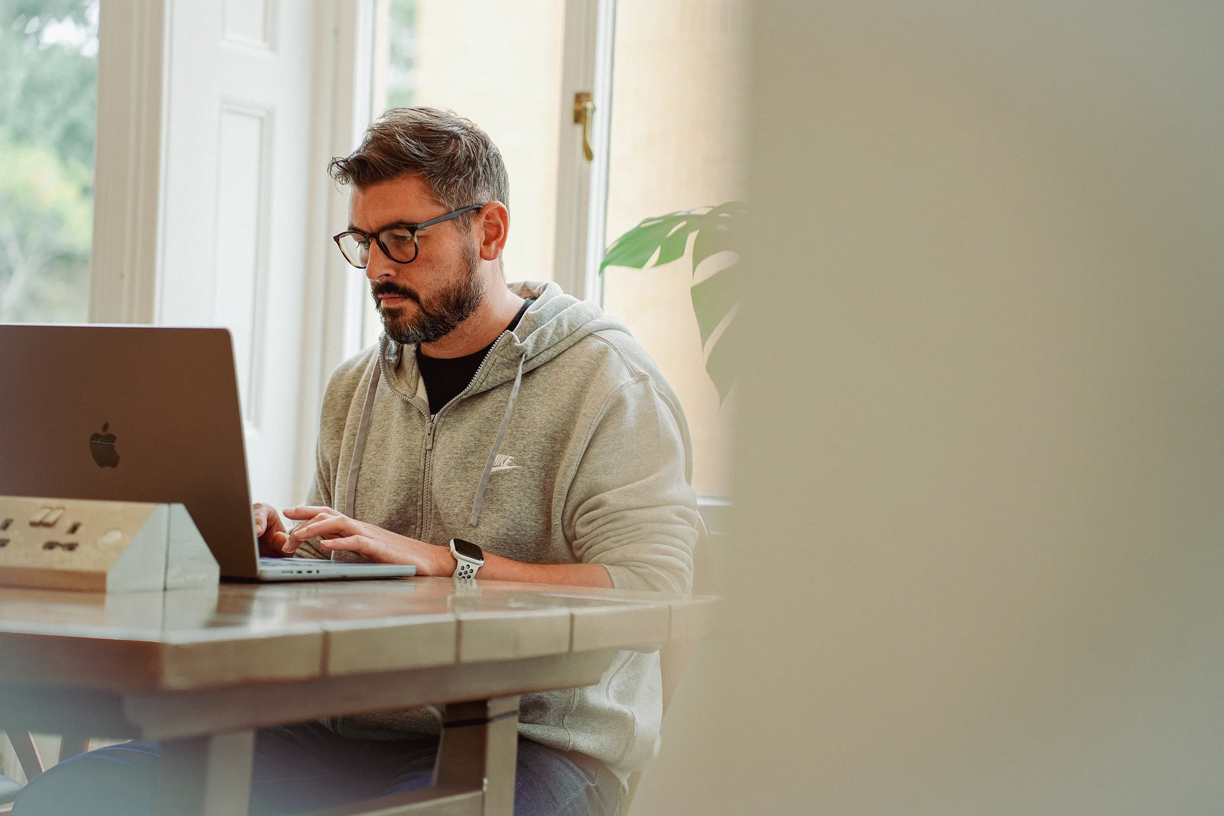 A man with glasses and a beard sitting at a table working on a laptop in a bright room with large windows and a potted plant.