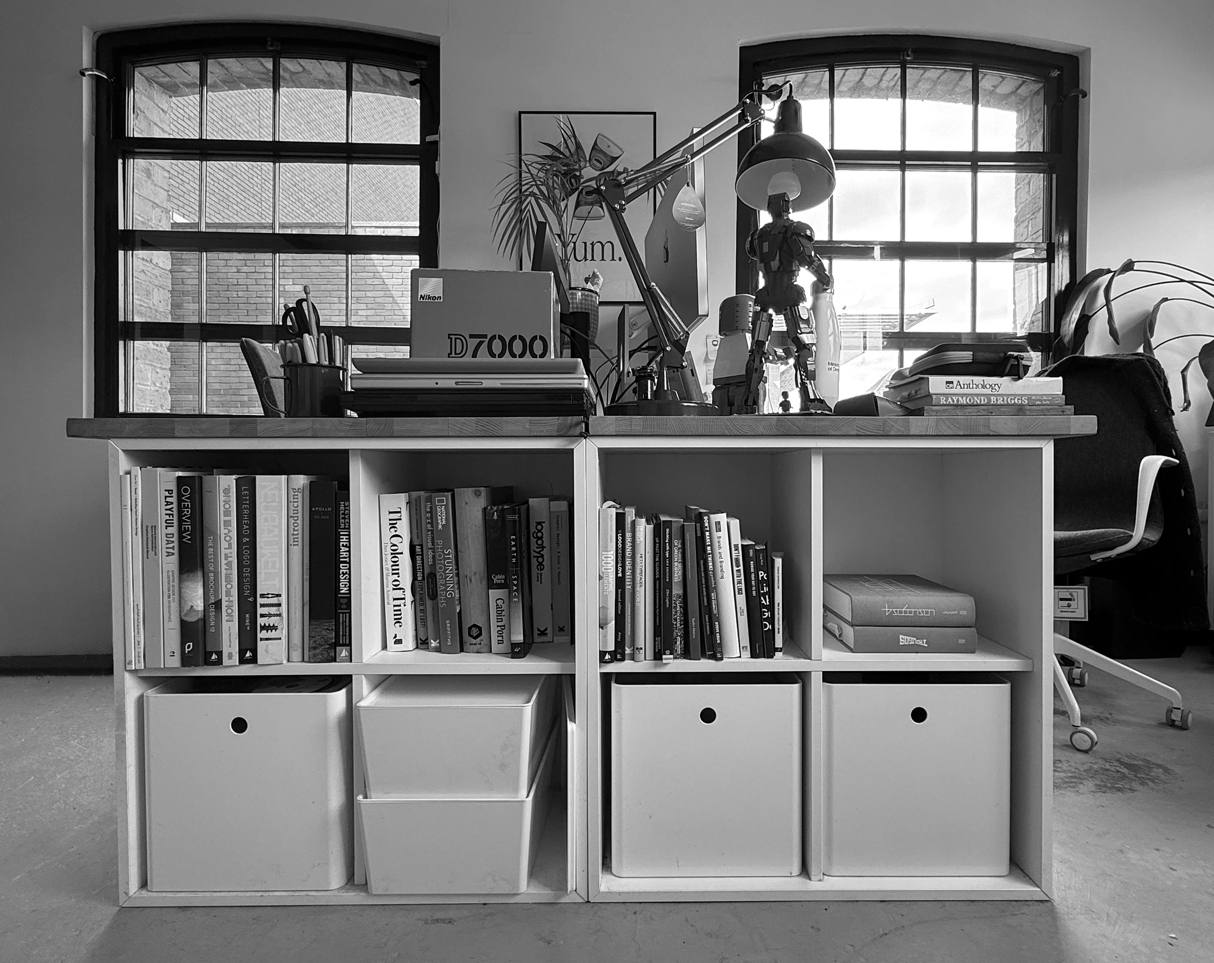 A white bookshelf filled with various books, with a desk behind it holding a Nikon D7000 camera, a lamp, a robot figurine, a stack of magazines, and some decorative items. There are two large windows in the background with brick walls visible outside.