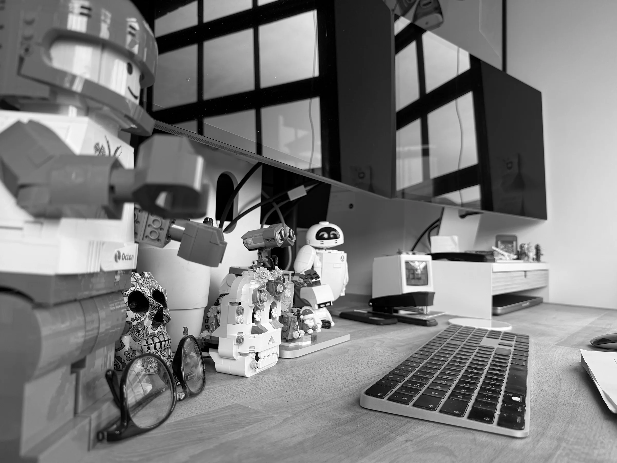 A workspace with a computer setup, including a keyboard and mouse, and various decorative items such as toy figurines, skull-shaped objects, and glasses, with shelves holding small figures and a monitor on the desk.