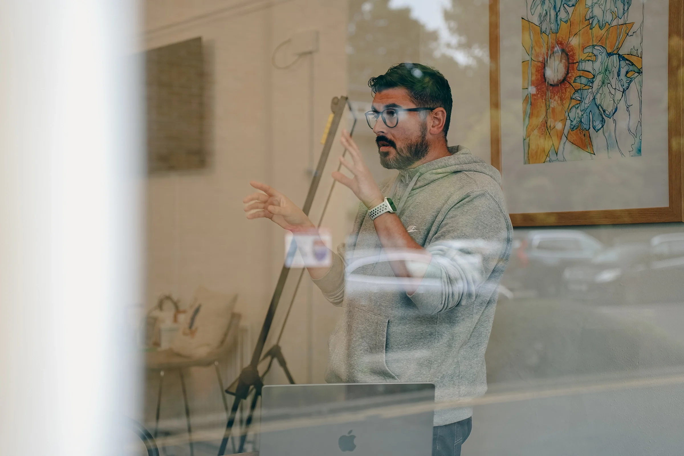 A man with glasses and a beard, wearing a gray hoodie, explaining something with hand gestures inside an art gallery with abstract paintings on the wall, viewed through a glass window.