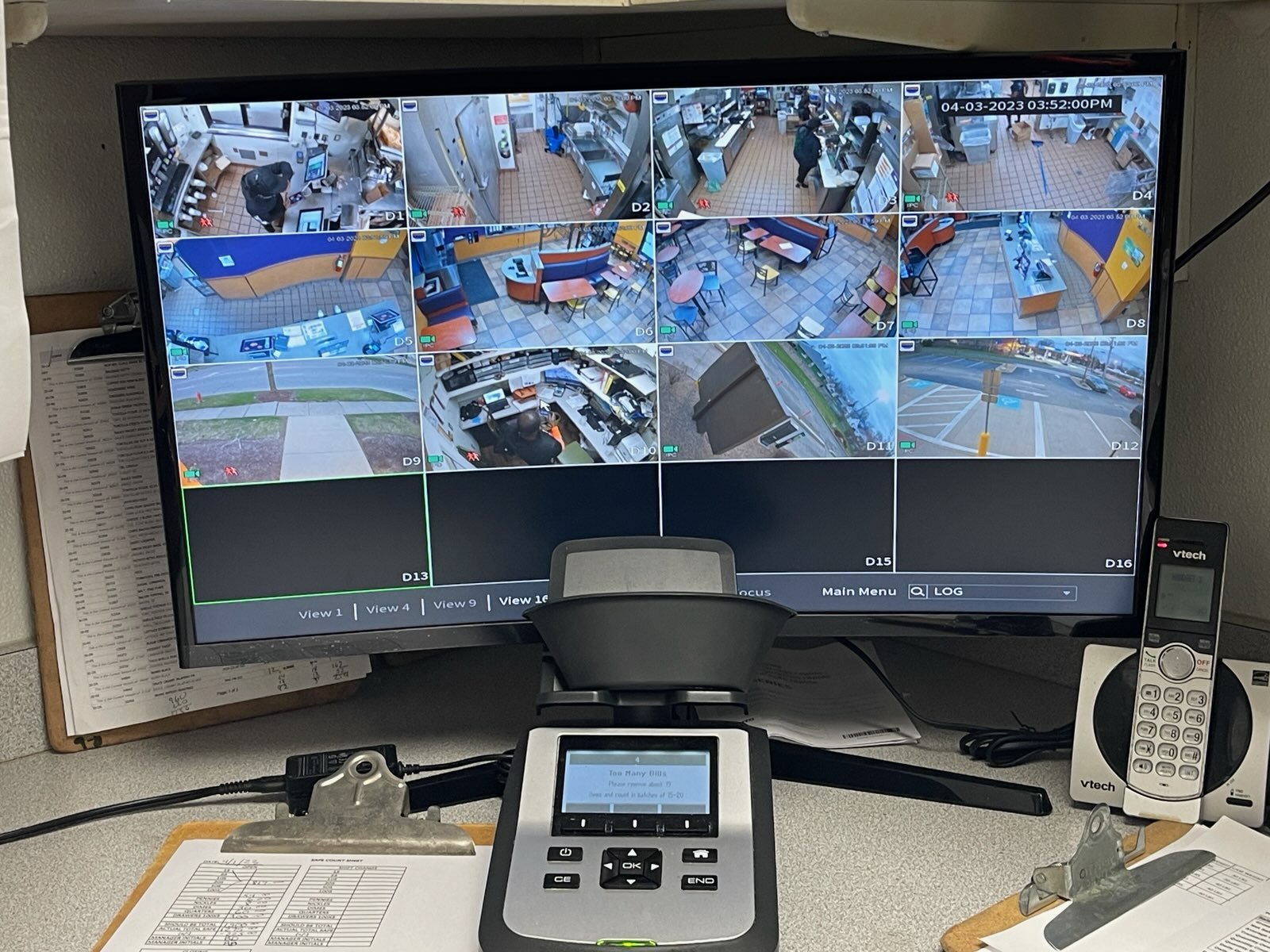 Security monitor displaying multiple camera feeds including a restaurant interior, outdoor sidewalk, and parking lot. Foreground shows a countertop with a VTech phone, a small keypad device, and documents. Office supplies and paper clip holder are al