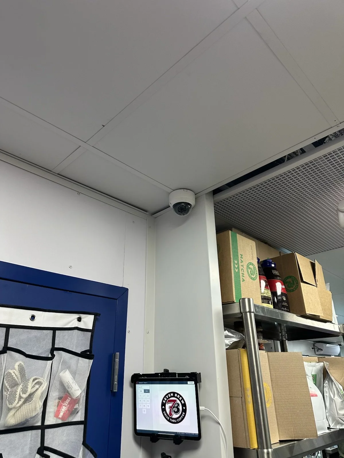 Security camera installed on the corner of a ceiling in a storage or back room with shelves, boxes, and beverages.