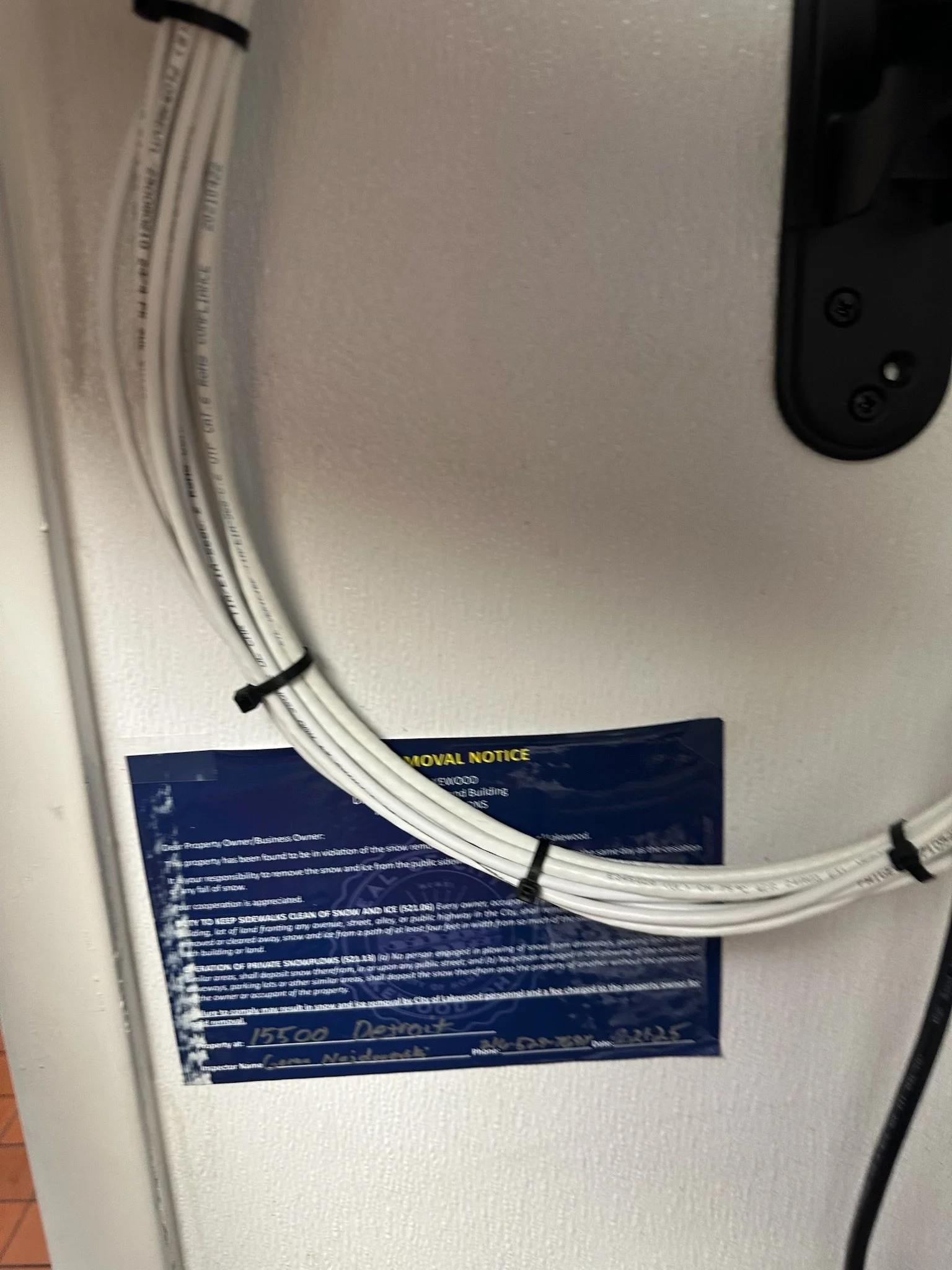 Close-up of a white cable bundle secured with black zip ties on a gray wall near a blue notice or sign with white and yellow text.