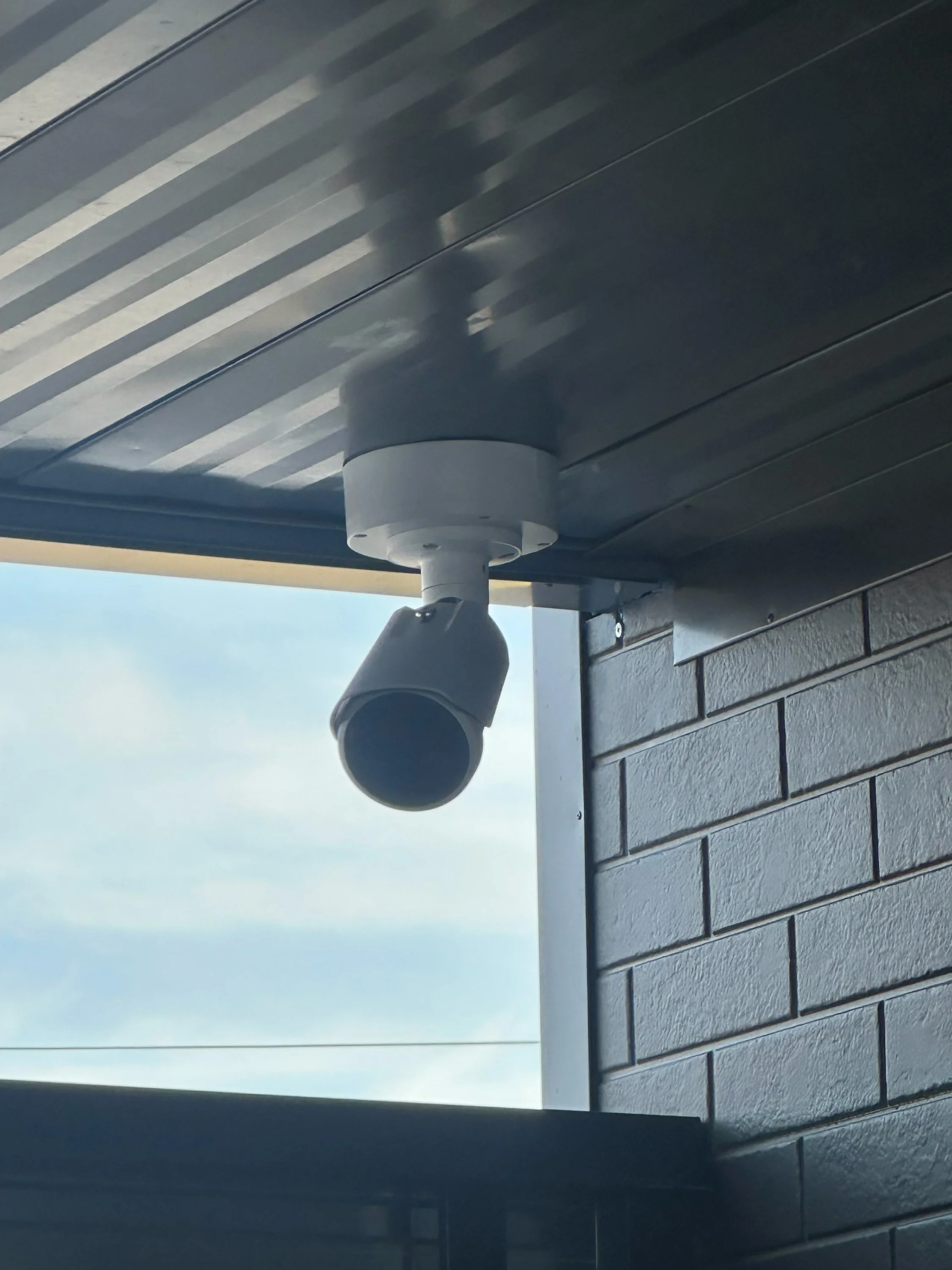 A security camera mounted on a building exterior beneath the roof overhang, with the sky visible in the background.