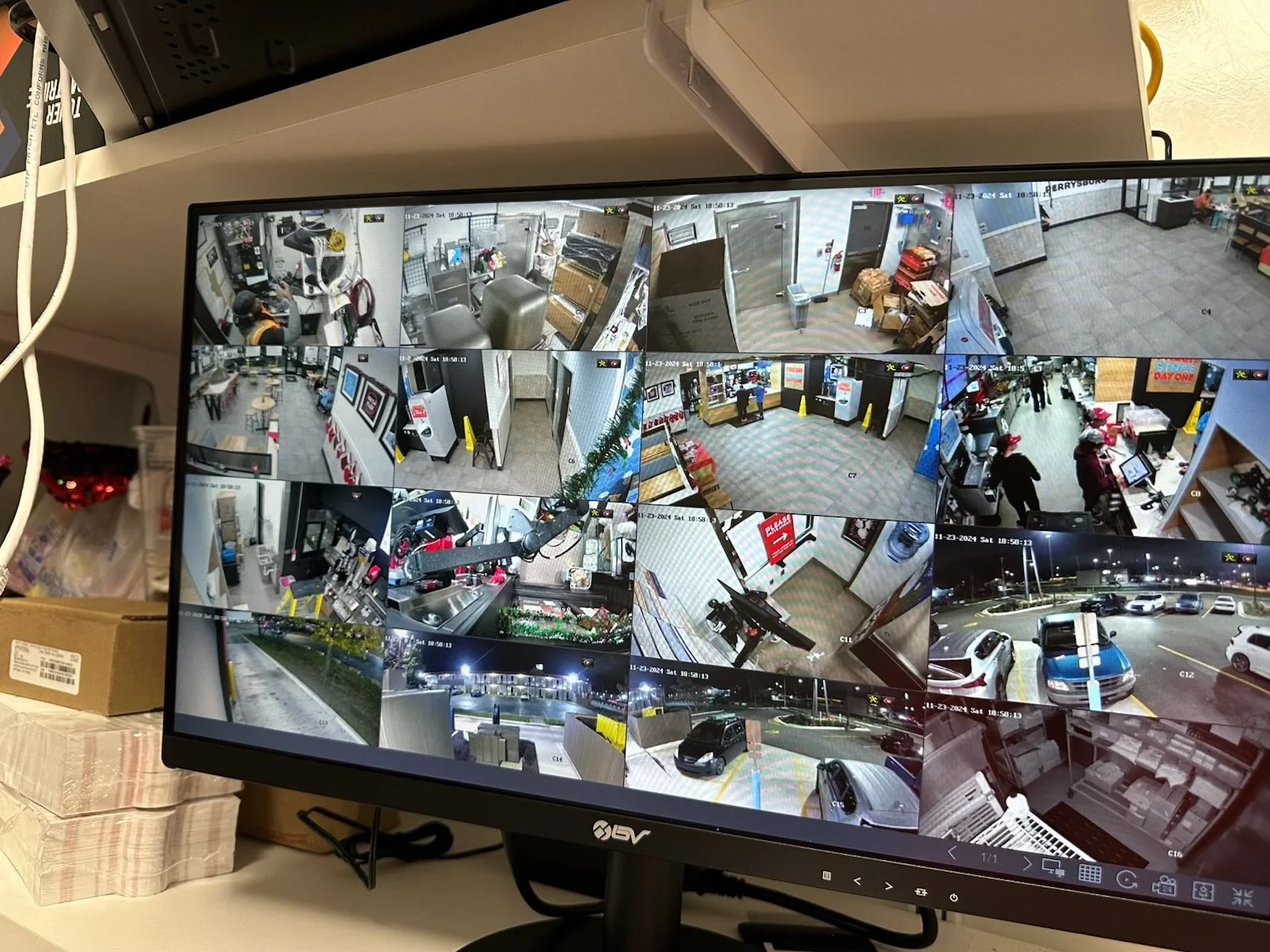 Security monitor displaying multiple CCTV camera feeds of various indoor and outdoor locations, including a cashier area, parking lot, and building entrances.