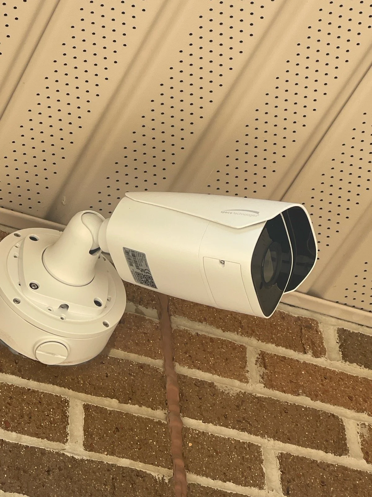 An outdoor security camera mounted on a brick wall, under beige perforated metal paneling.