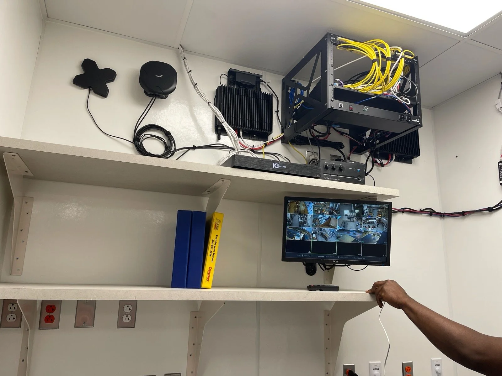 Room with electronic equipment including a surveillance monitor, a rack with yellow cables, two black heat sinks, a hand plugging in a cable, and a shelf with boxes and second shelf with power outlets.