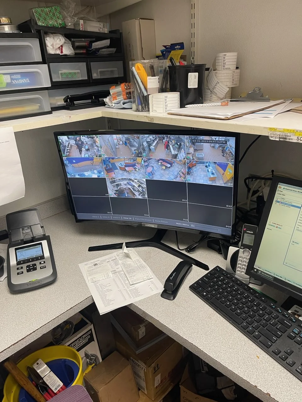 Office security monitoring station with a monitor displaying multiple surveillance camera feeds, a computer, a desk phone, and various office supplies.
