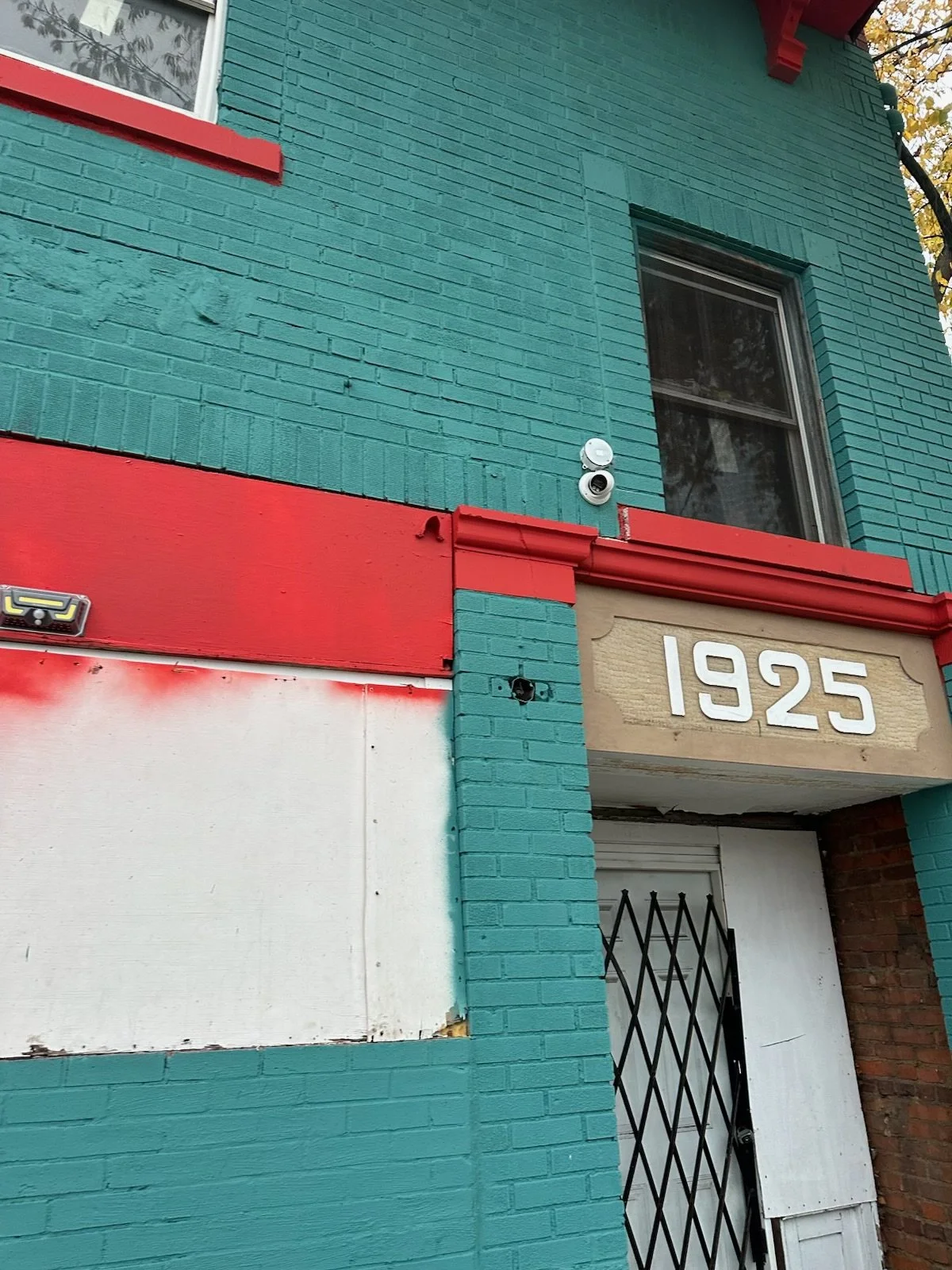 Facade of a building painted turquoise with red accents, displaying the address '1925' on a sign over the doorway. The building has a window, security camera, and some boarded-up areas.