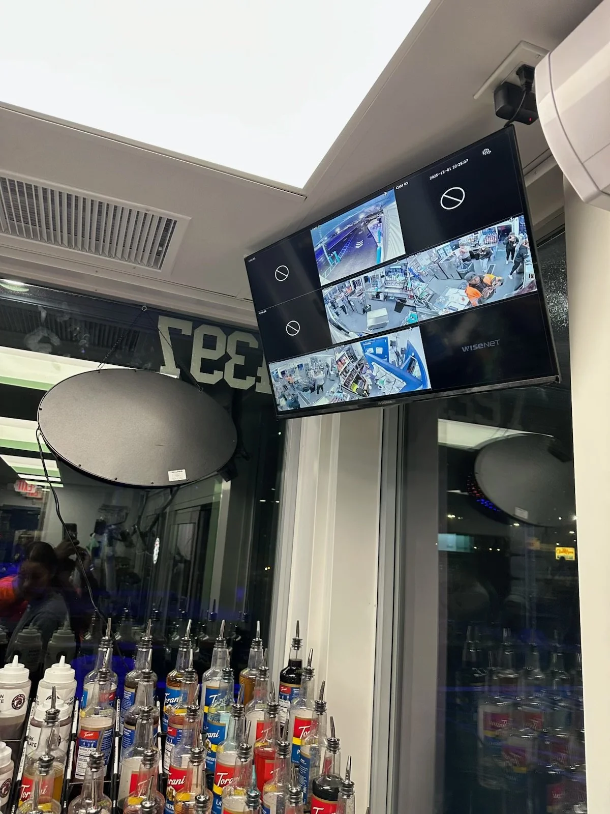 Security camera monitors displaying surveillance footage inside a store, with bottles of condiments in the foreground and a reflection of people in the window.
