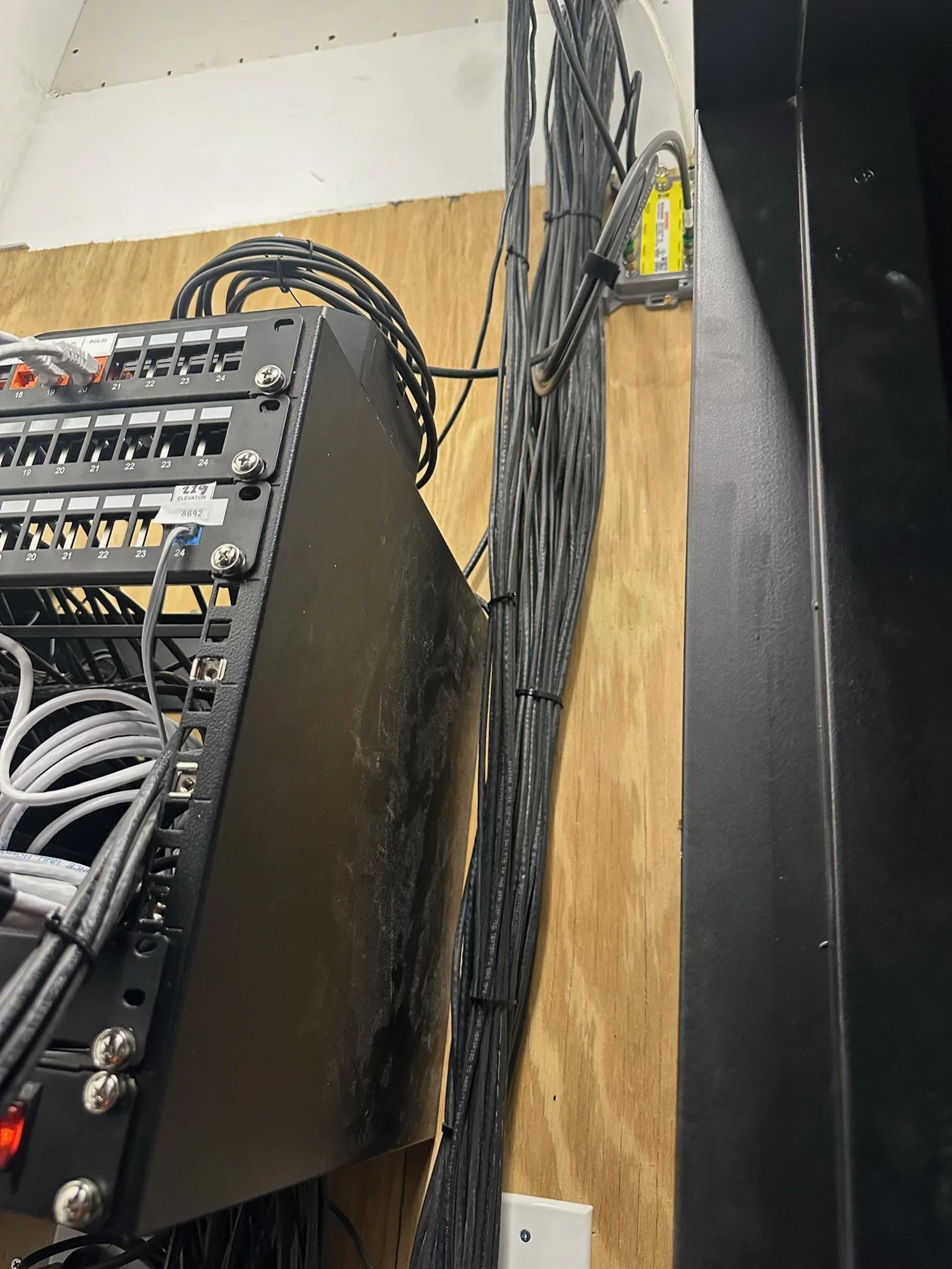 Close-up of a black electronic rack with circuit boards, cables, and connectors, mounted on a wooden surface. Some cables are bundled and secured with cable ties. An electrical outlet is visible at the bottom.