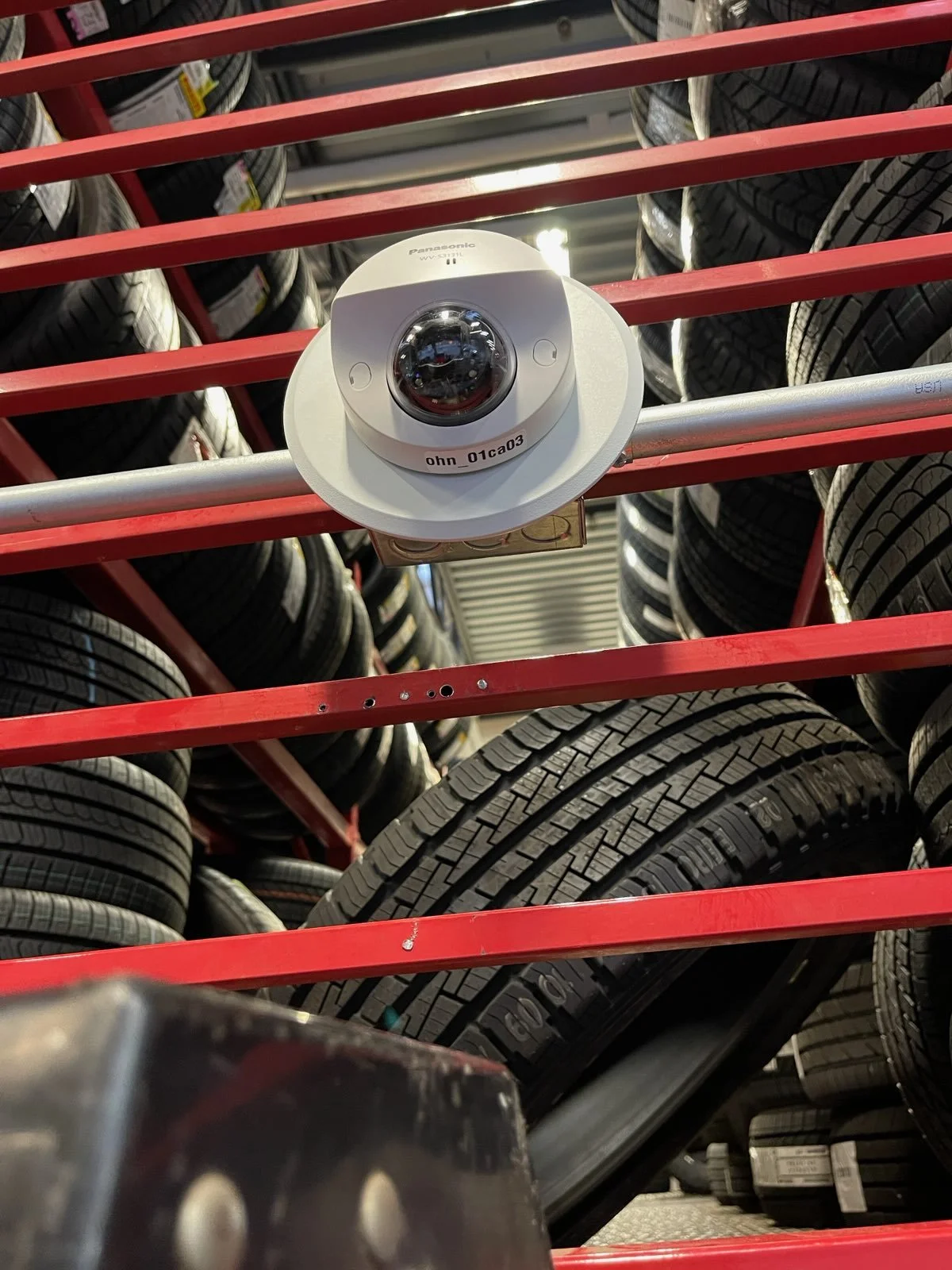 A security camera mounted on a metal bar inside a tire store, with stacks of tires in the background.