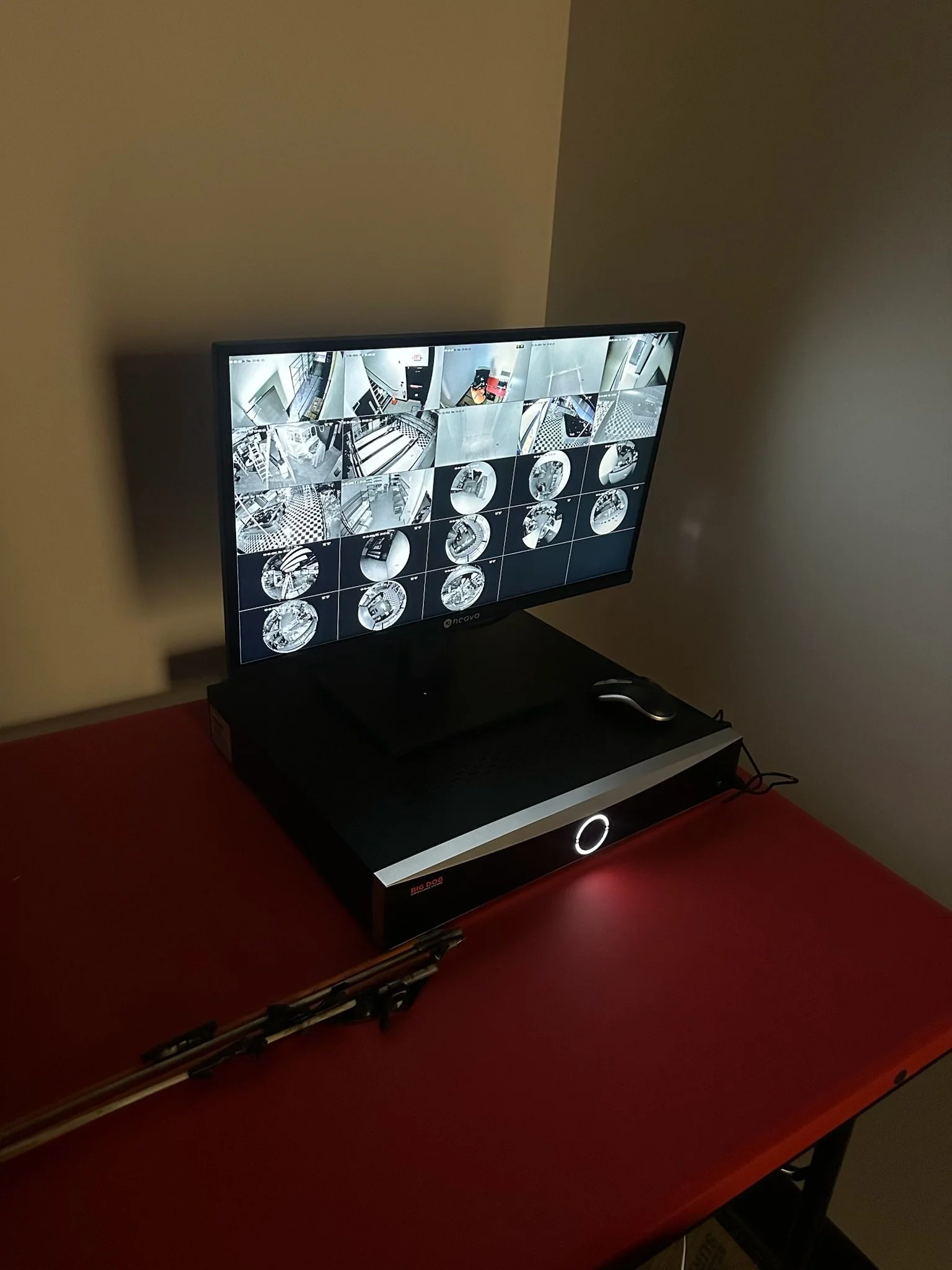 A computer monitor displaying multiple surveillance camera feeds mounted on a black desk with a red surface, next to a microphone stand.