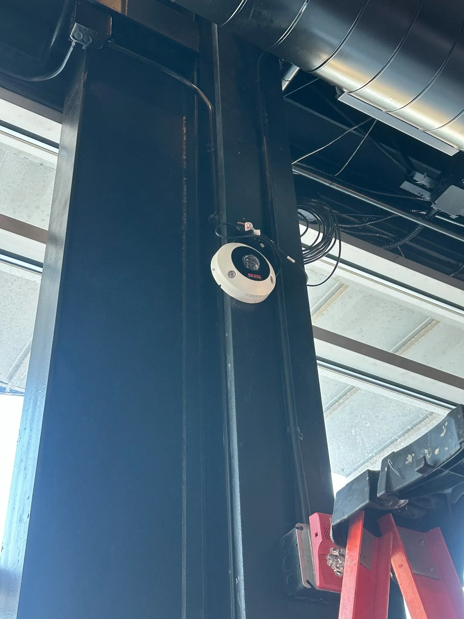 A security camera mounted on a black metal support beam inside a building with a glass ceiling.
