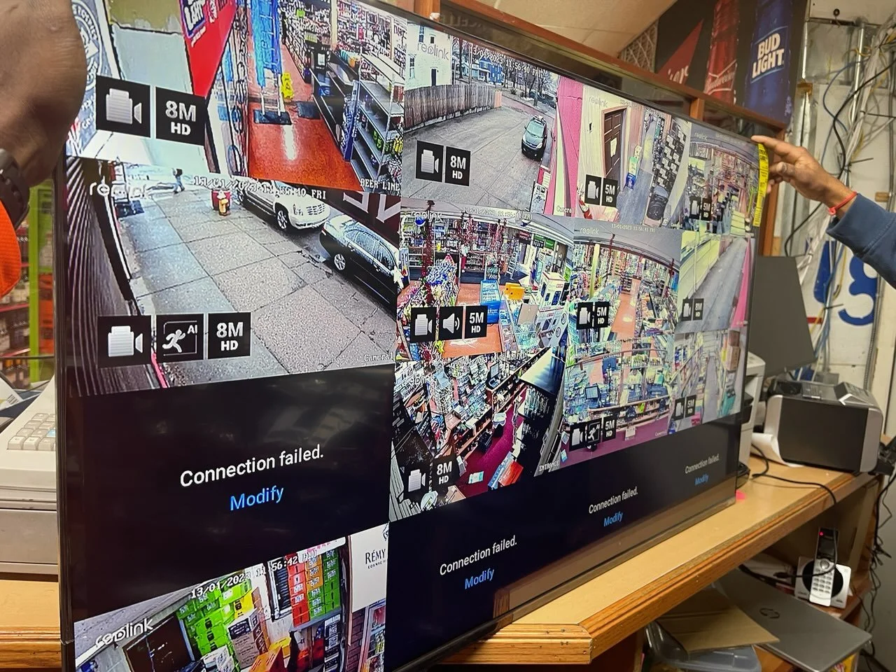 A large flat screen monitor displaying multiple security camera feeds in a store, with some showing connection failed messages. The monitor is on a wooden desk with office equipment around.