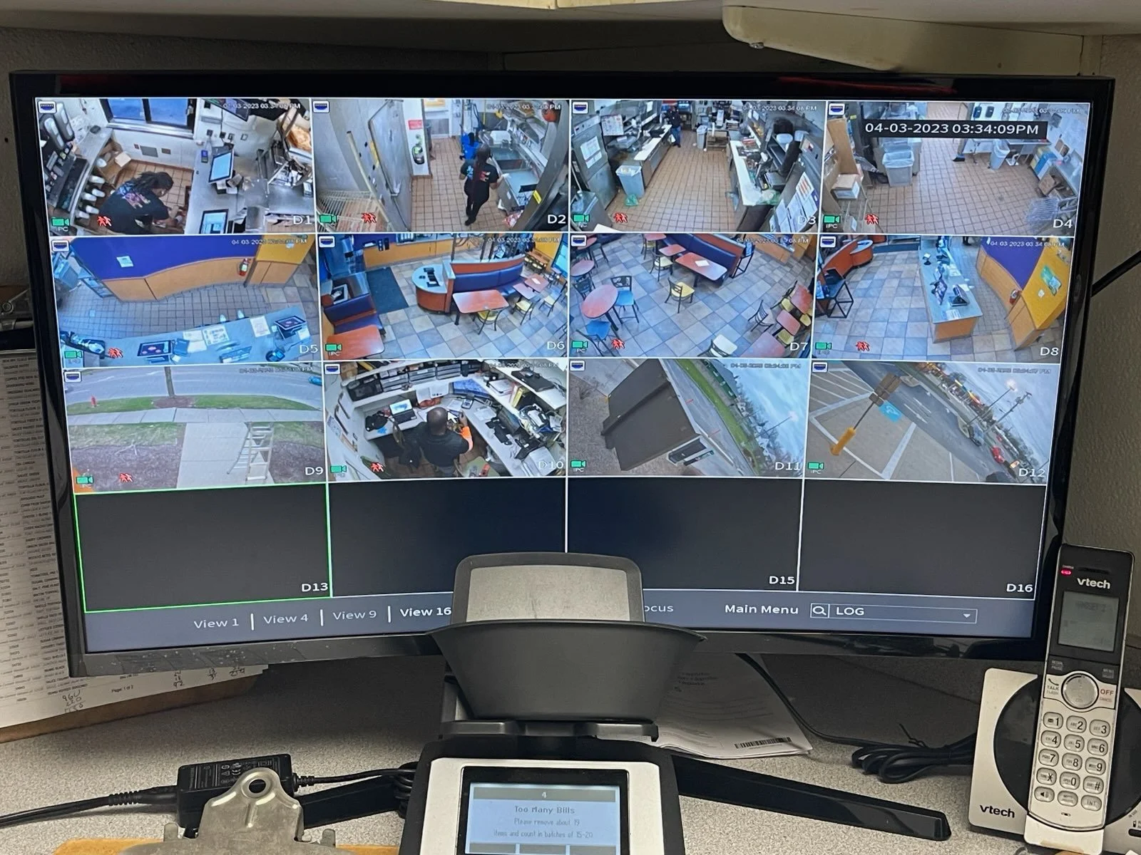 Multiple surveillance camera feeds on a monitor showing various scenes inside and outside a restaurant or fast-food establishment, including inside the kitchen, dining area, and outside parking lot.