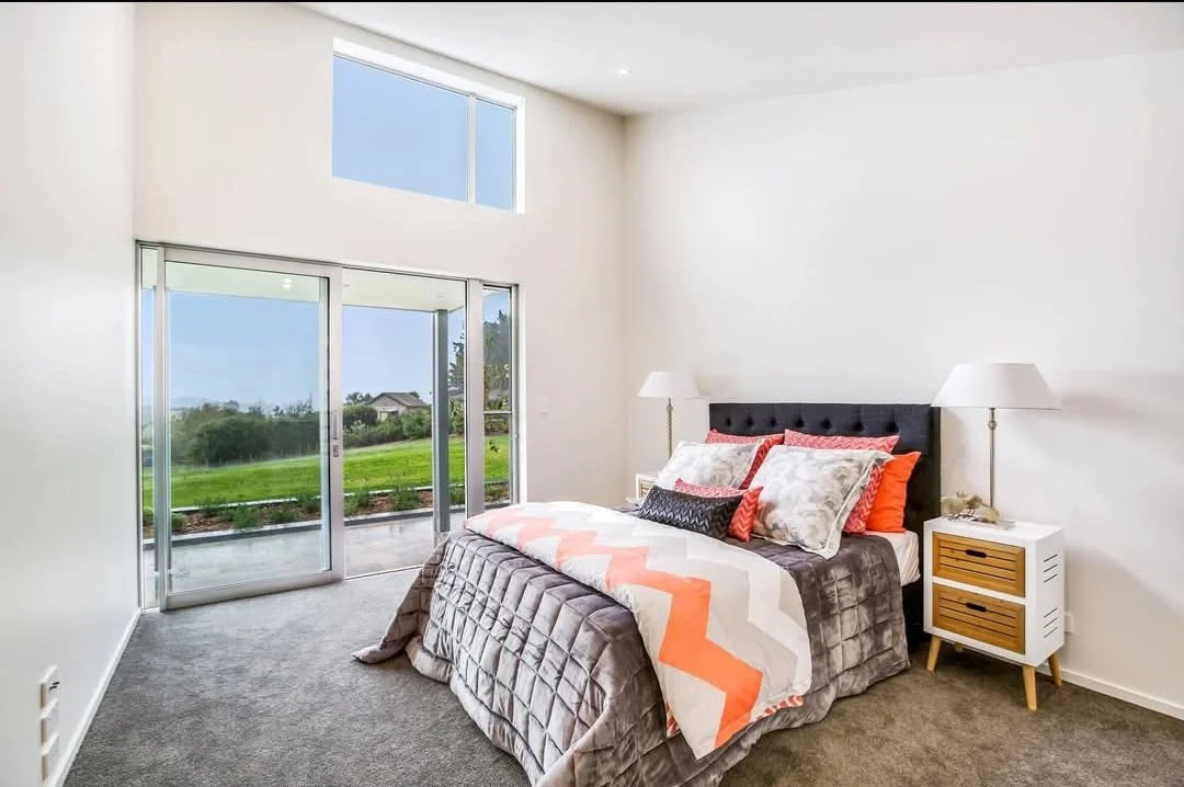 A modern bedroom with white walls, a large bed with a black headboard, assorted pillows, and a geometric-patterned blanket. Two white nightstands with wooden drawers and table lamps are on either side of the bed. Sliding glass doors and windows provide a view of green grass and a clear sky.