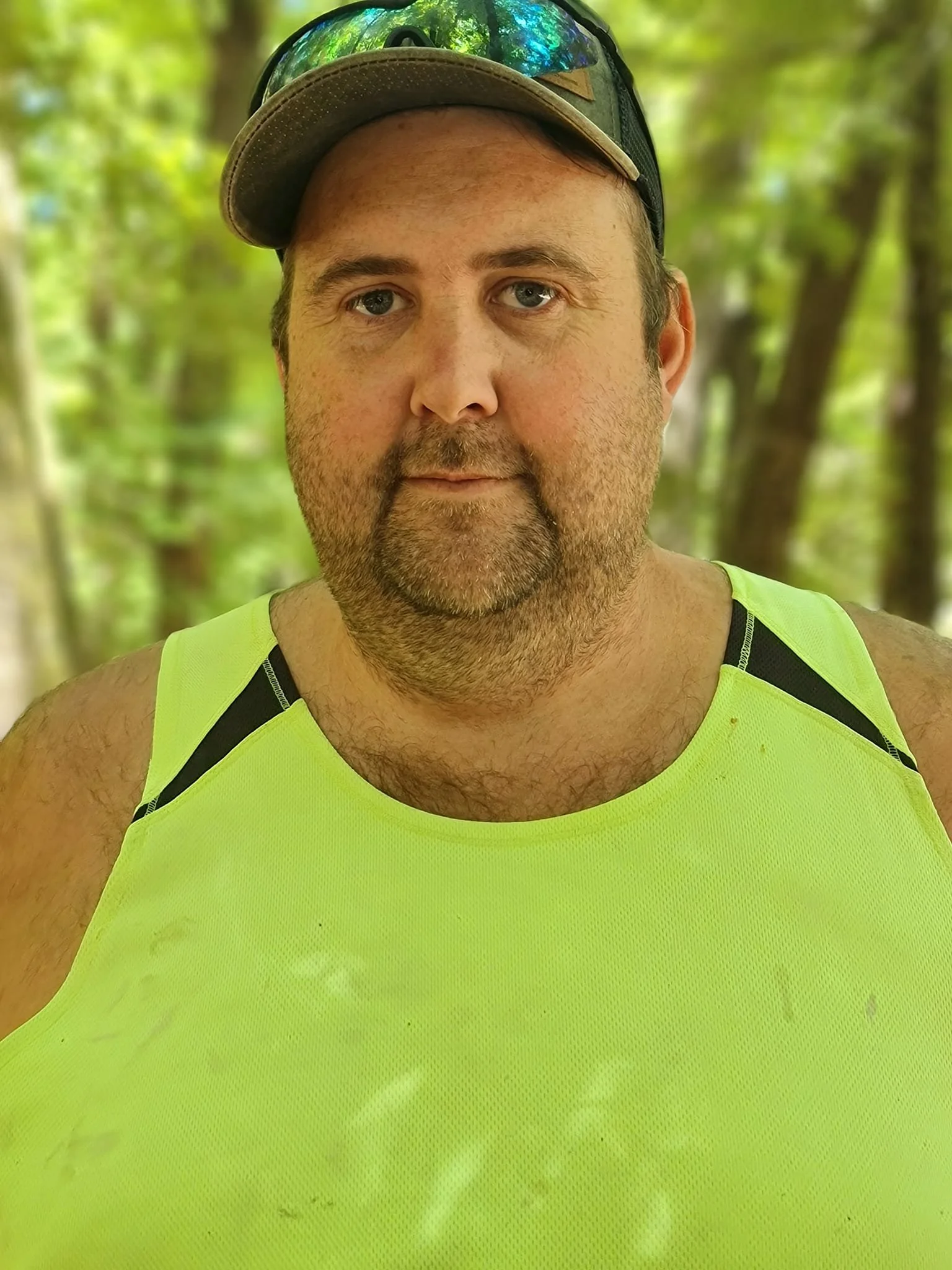 A man wearing a tan baseball cap with sunglasses on top, a bright yellow sleeveless athletic shirt, and a black tank top underneath, standing outdoors in a wooded area.