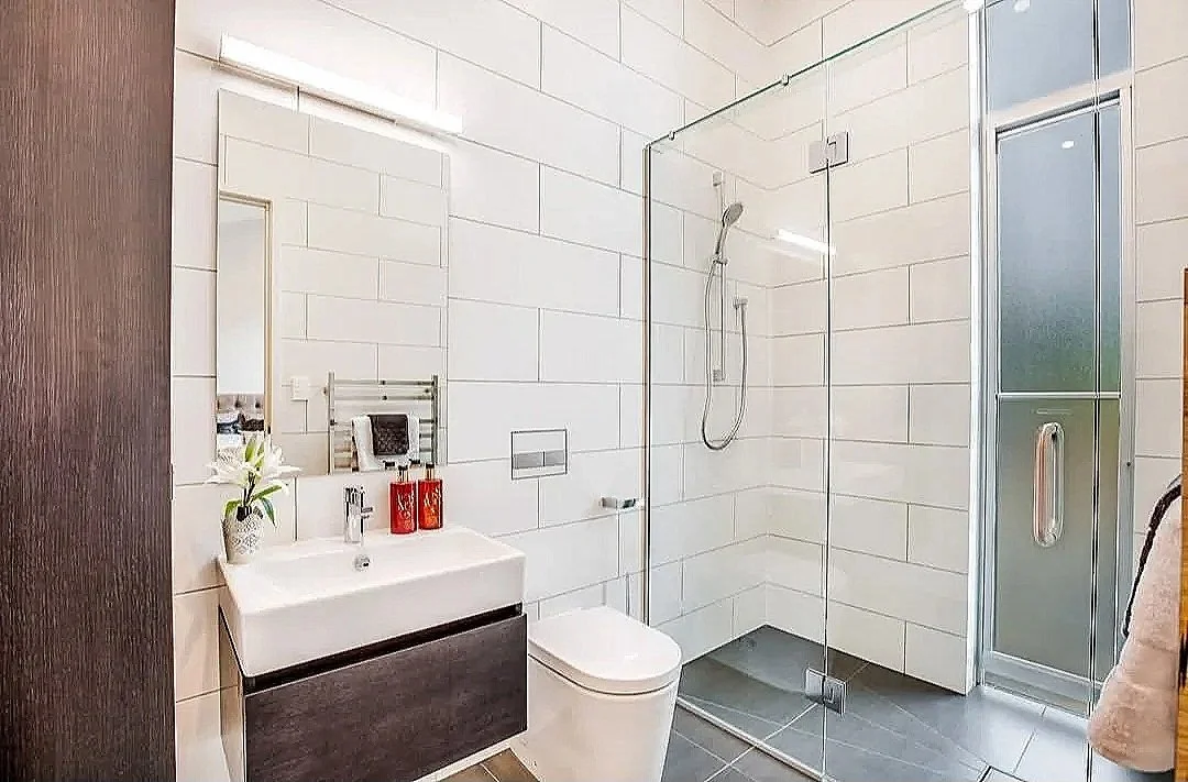 Modern bathroom alteration with white tiled walls, glass-enclosed shower, white toilet, and wall-mounted sink.