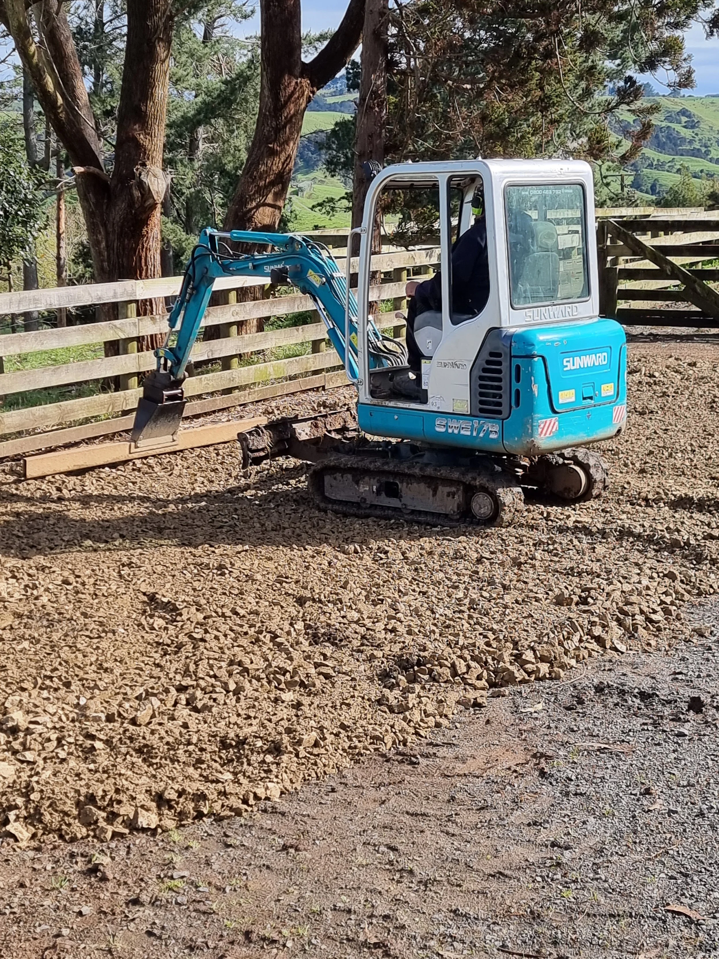 A small blue and white excavator working on a dirt area near a wooden fence and trees with rolling green hills in the background.