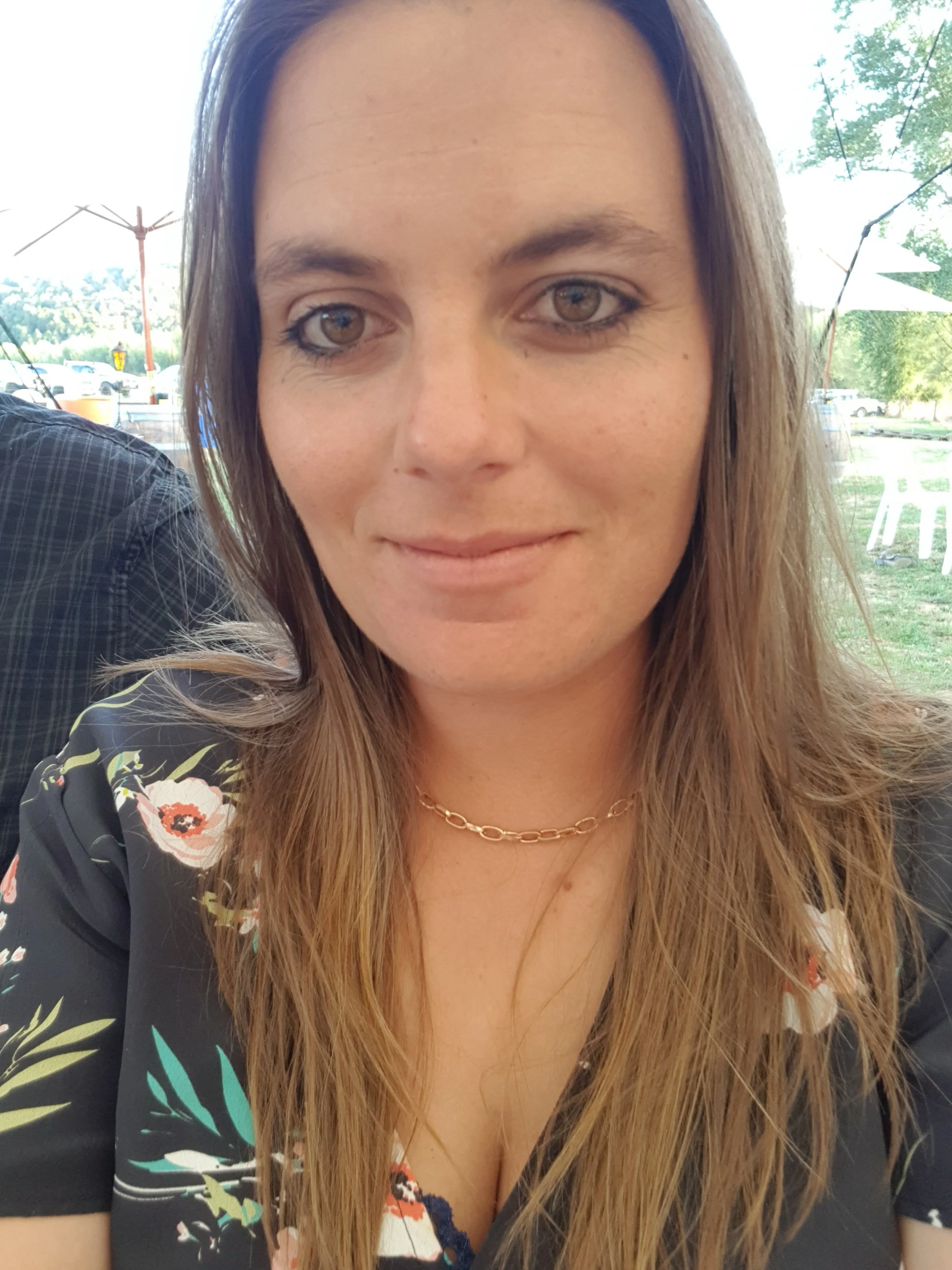 A woman with long brown hair, hazel eyes, wearing a gold necklace and a floral black top, outdoors near tables and umbrellas, with trees and parked cars in the background.