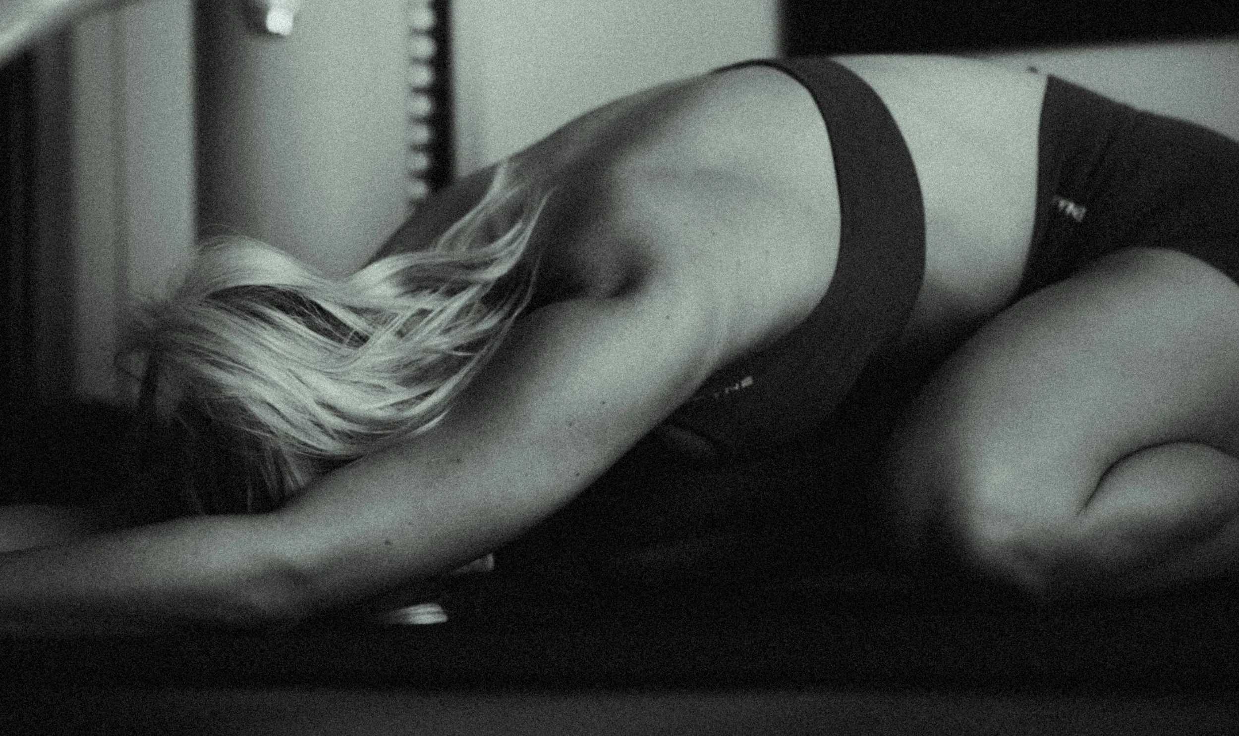 A woman with long wavy hair lying on her stomach on a bed or couch, wearing a black sleeveless top, with her back to the camera in a black and white photograph.