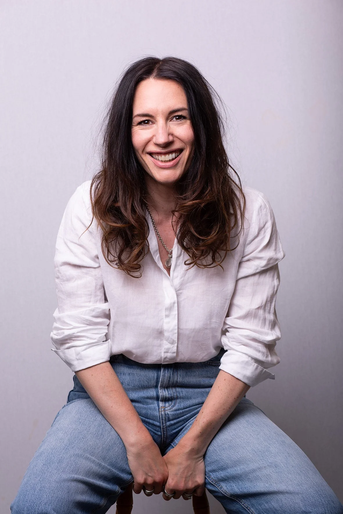 Woman with dark shoulder-length hair, smiling, wearing a white shirt and light blue jeans, sitting against a light gray background.