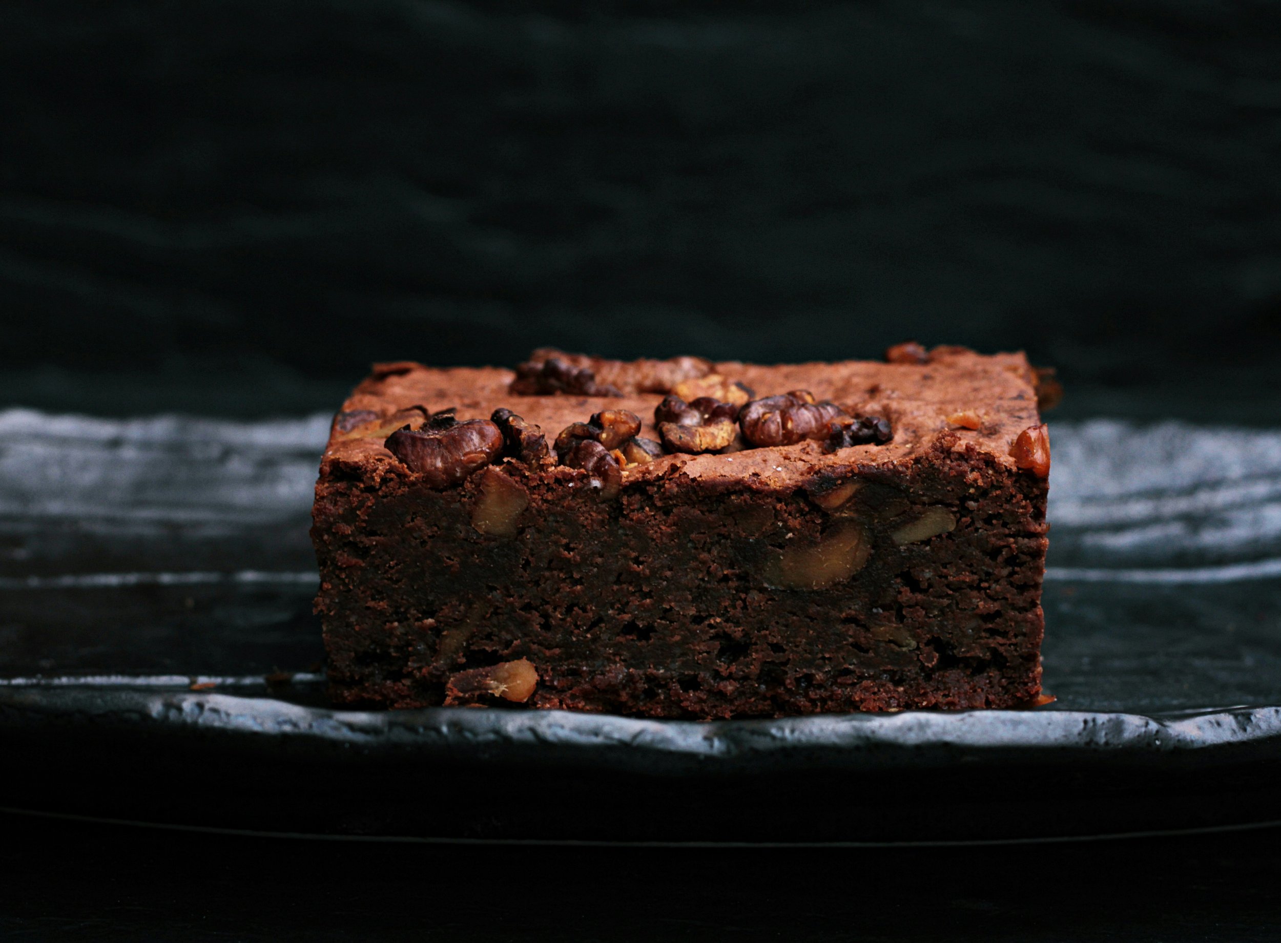 A close-up of a chocolate brownie topped with nuts, placed on a reflective surface with a dark background.
