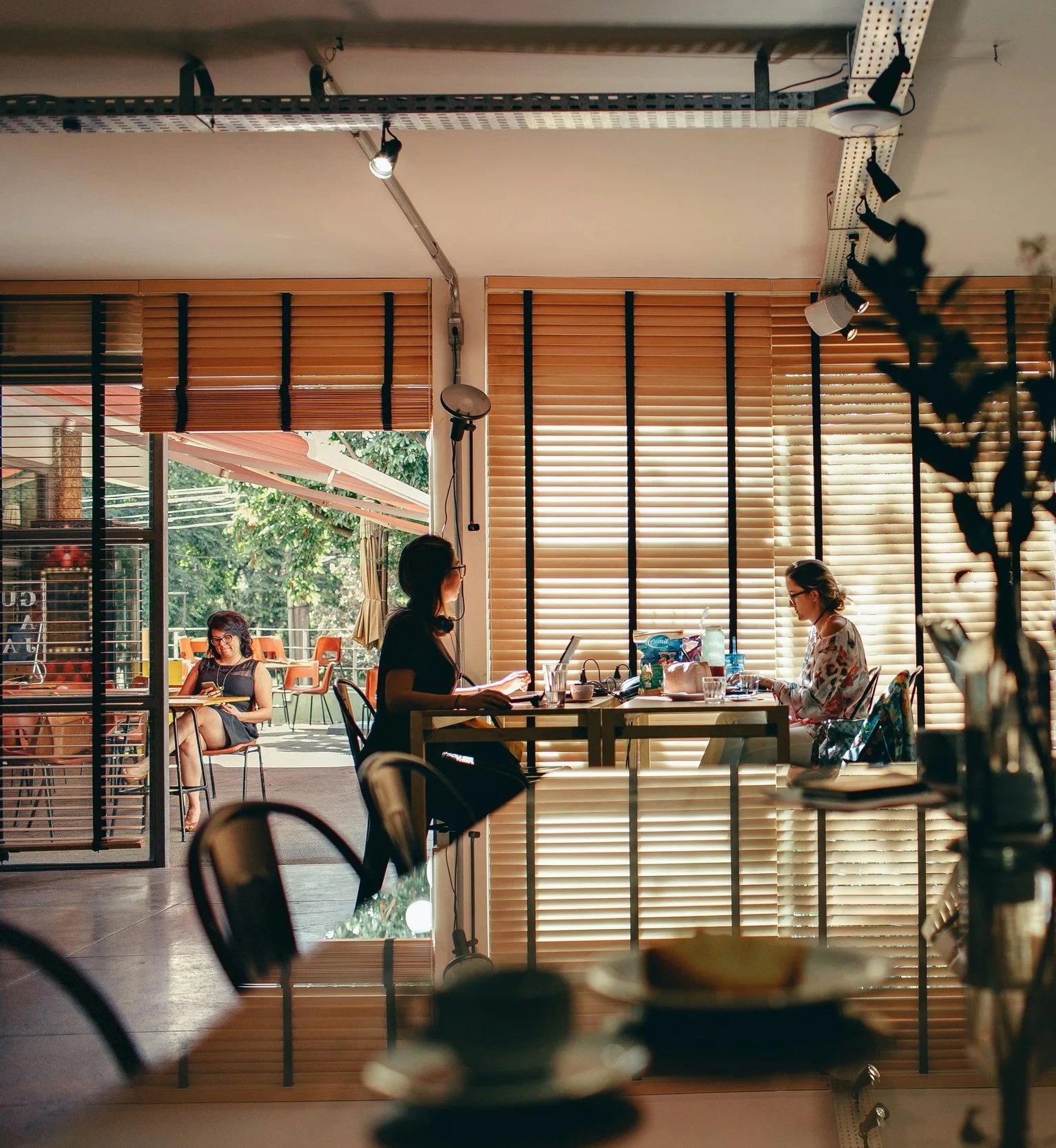 Interior of a cafe with three women using laptops and tablets, wooden blinds on windows, and outside patio with woman sitting and reading.