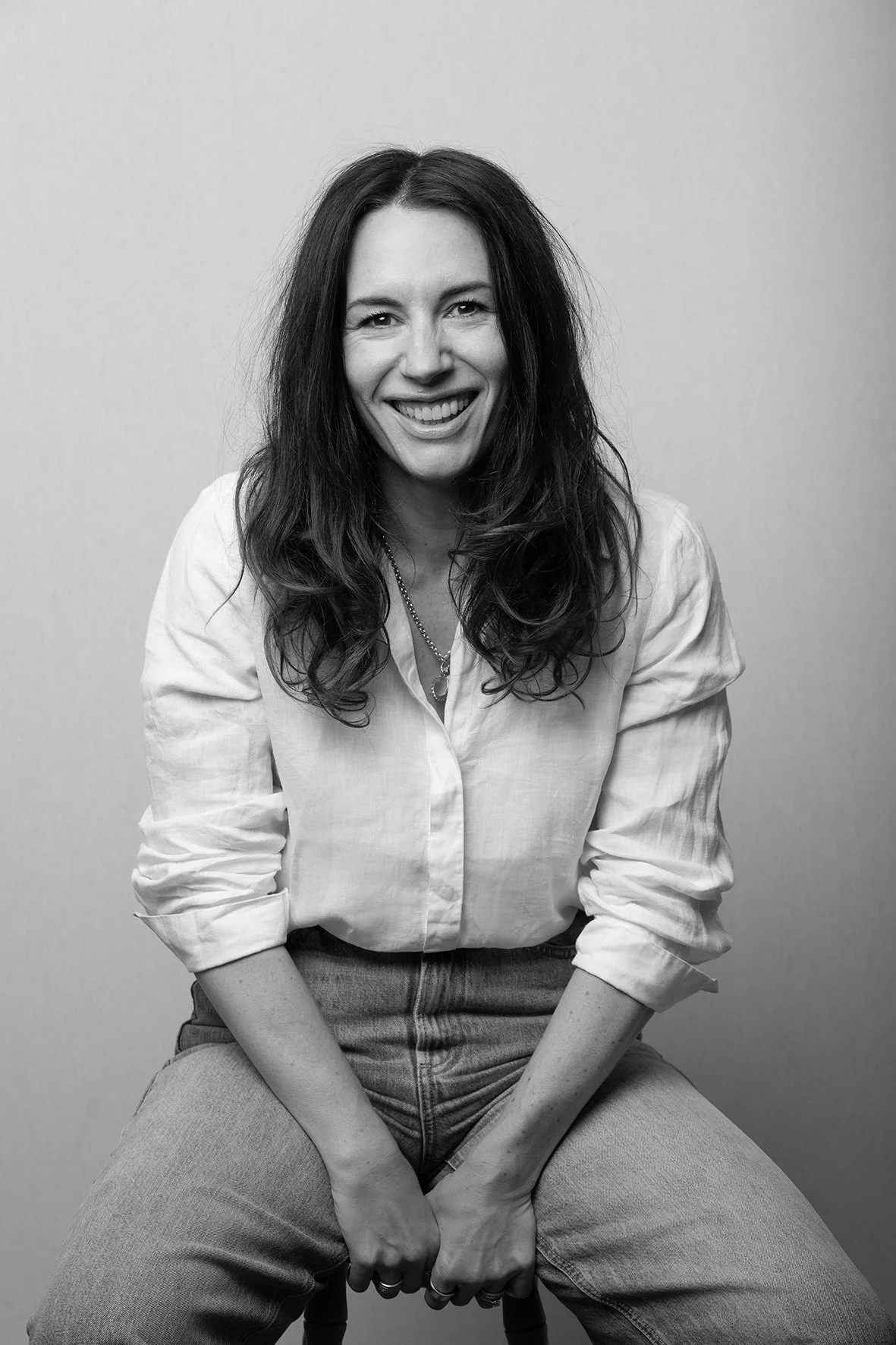 Black and white portrait of a woman with long wavy hair smiling, sitting with legs apart and hands clasped, wearing a white shirt and jeans, against a plain background.
