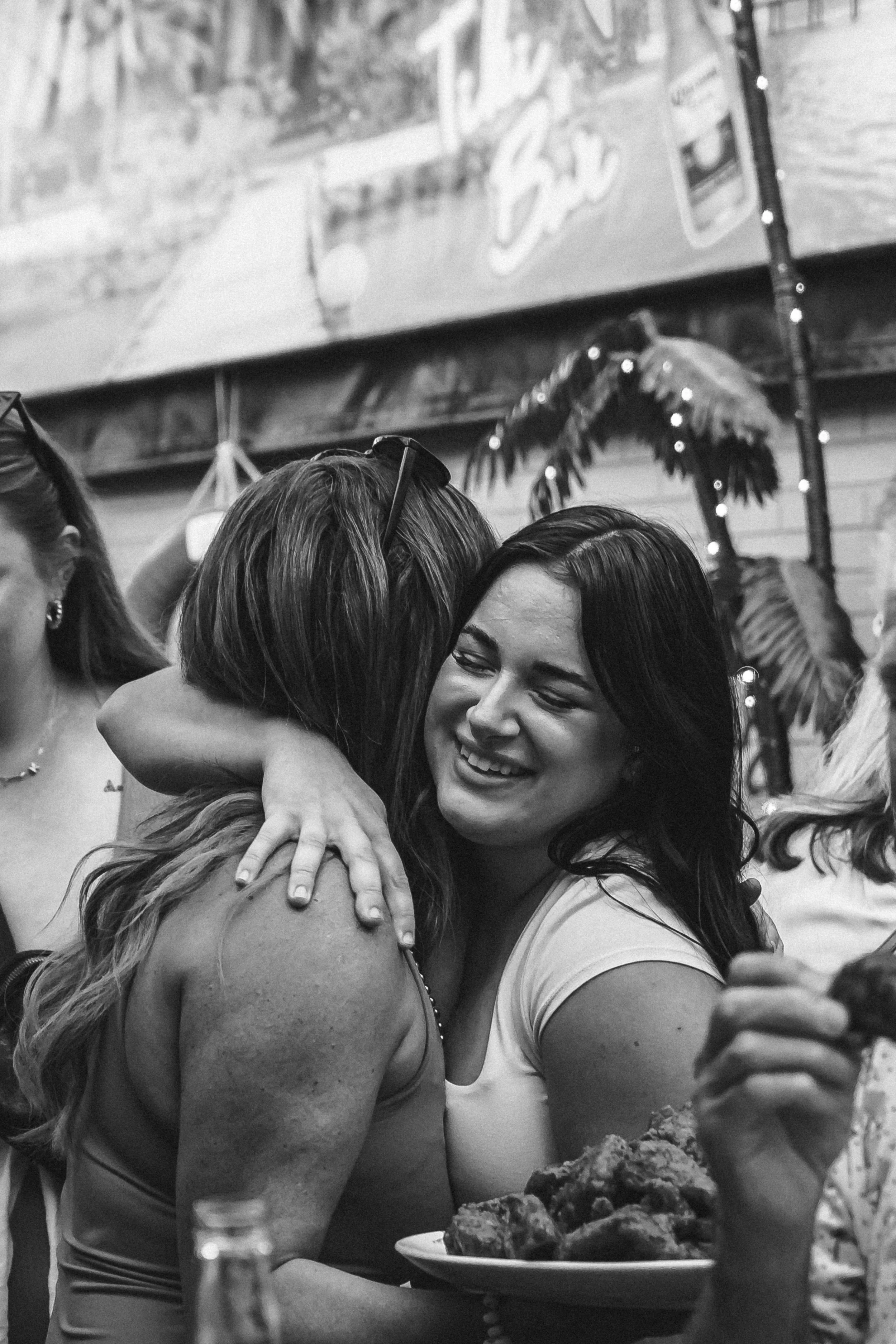 Two women embracing at a social gathering, smiling and holding a plate of food, with other people and decorations visible in the background.