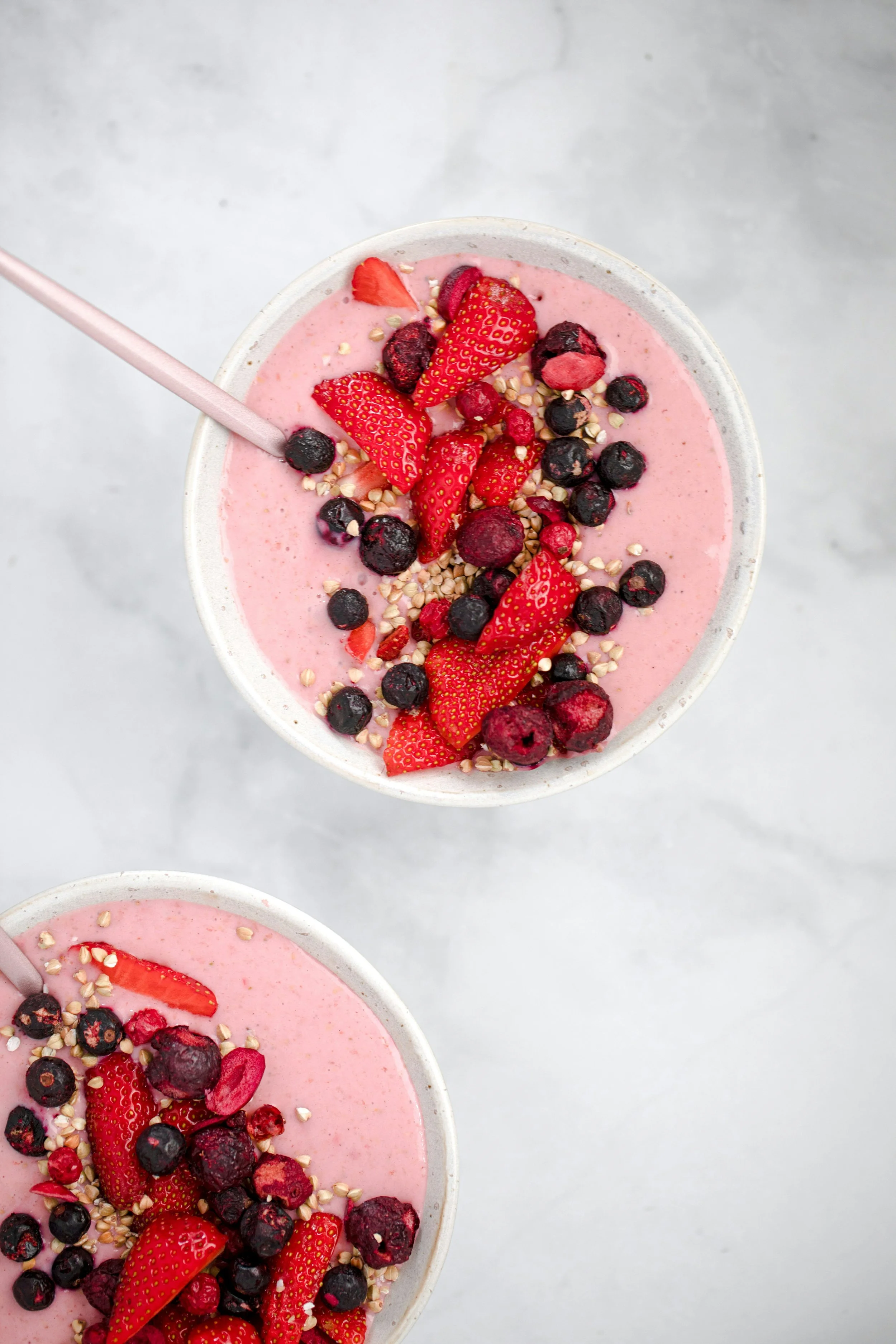 A cup of pink smoothie topped with sliced strawberries, blueberries, red berries, chopped nuts, and a pink straw, on a gray surface.