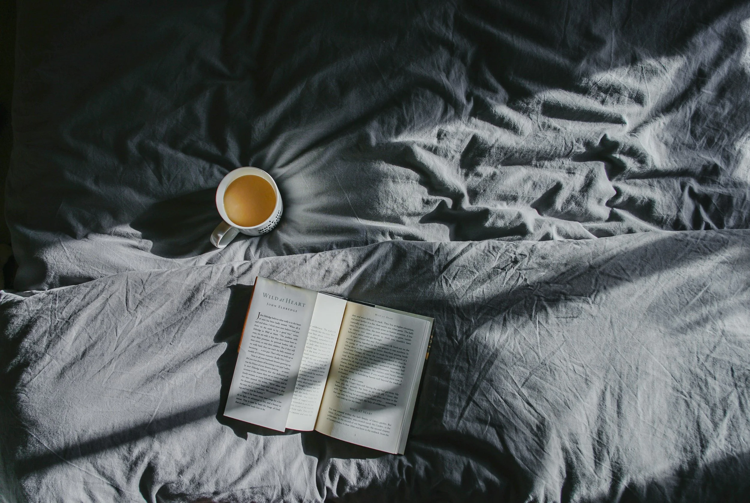 A white mug filled with coffee resting on a rumpled gray bedsheet, with an open paperback book nearby and sunlight casting shadows.