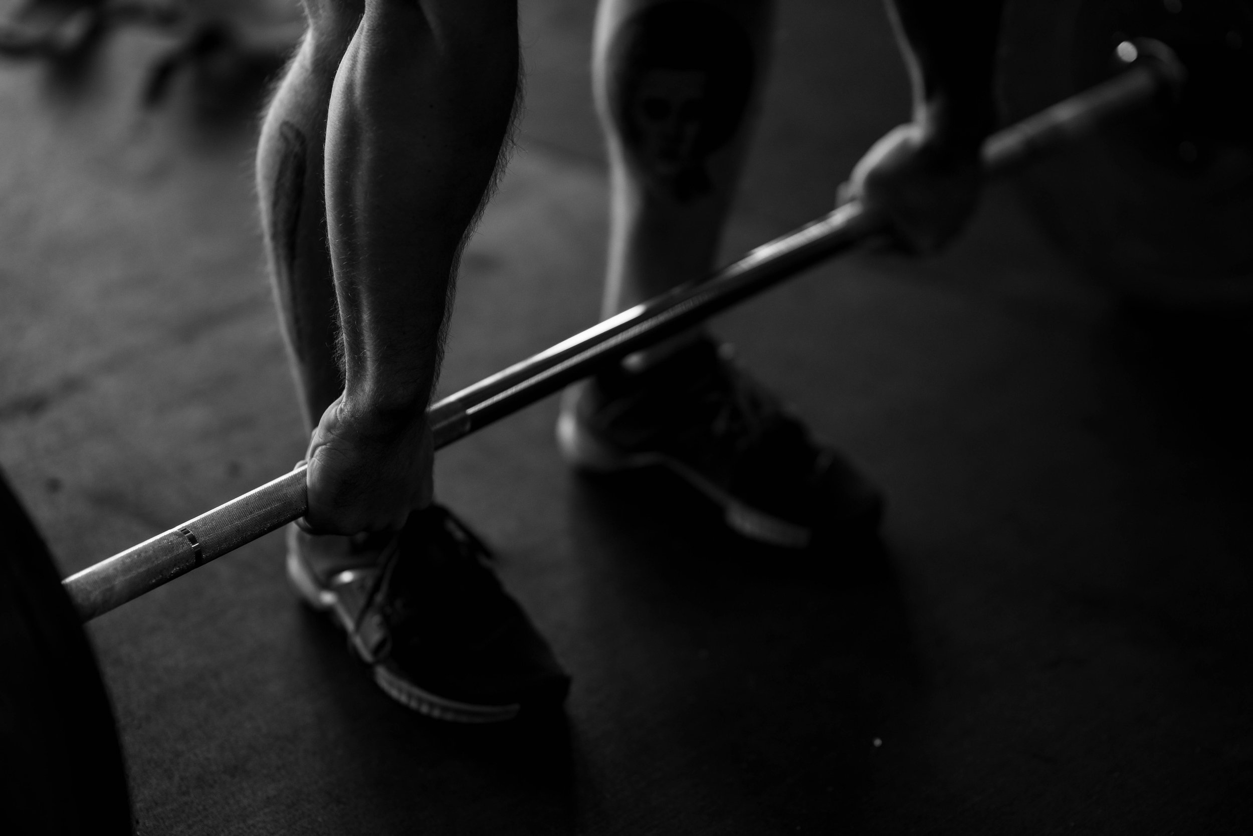 Black and white photo of a person lifting a barbell in a gym with a focus on their arm and legs.