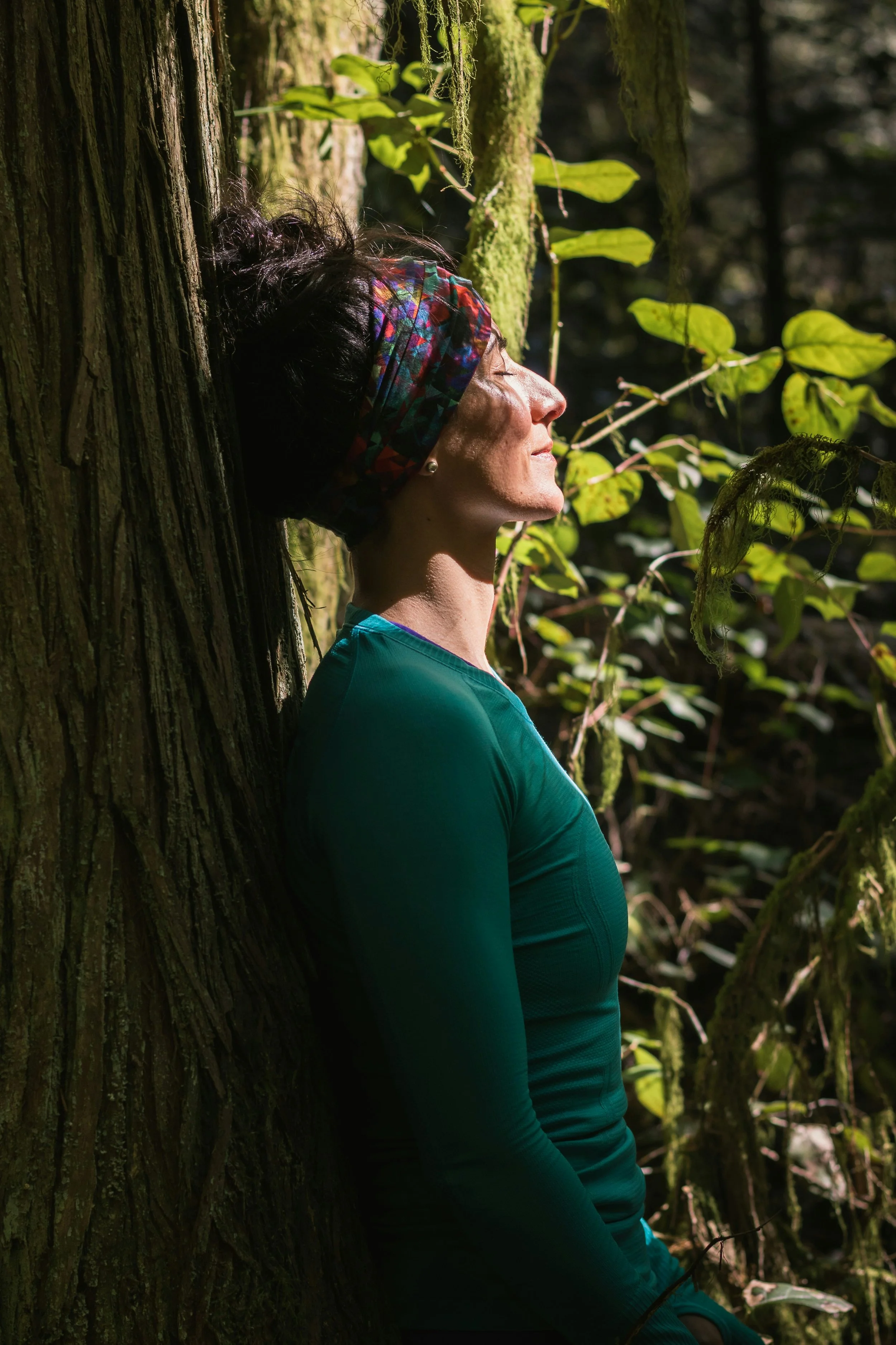 A woman with dark curly hair, wearing a colorful headband and a teal shirt, leans against a moss-covered tree in a forest with sunlight filtering through the leaves.