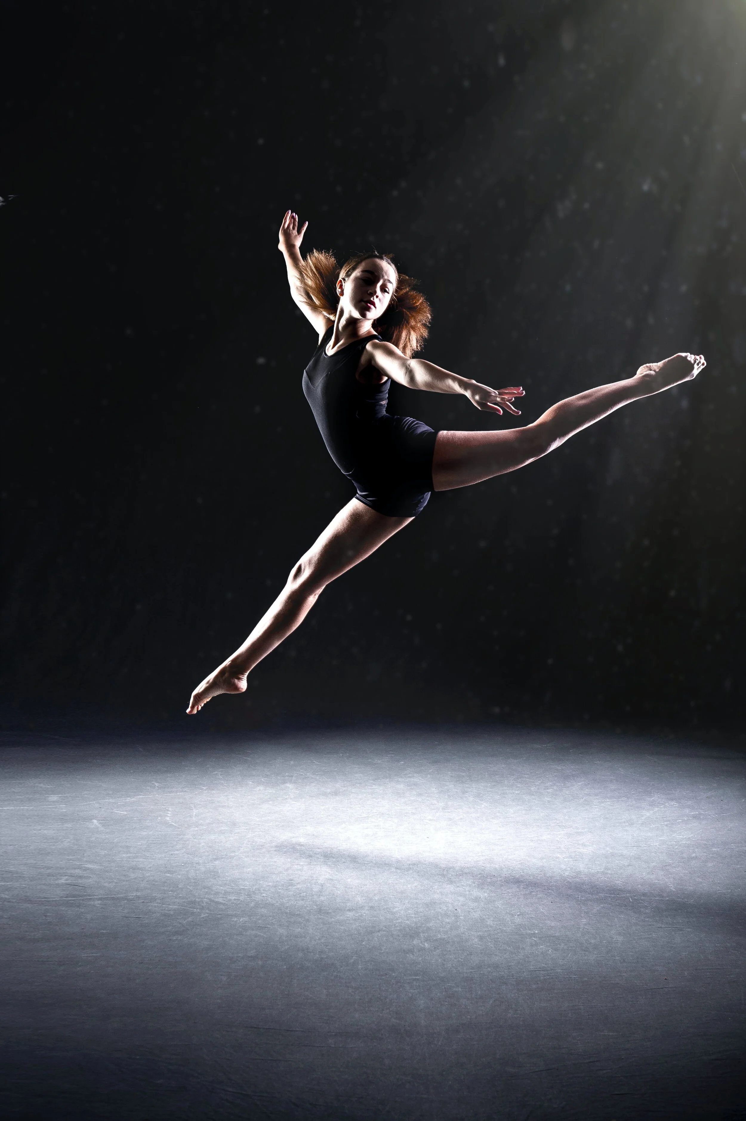 A female ballet dancer in a leap performance, wearing a black leotard, in a dark studio with spotlight and dust particles in the air.
