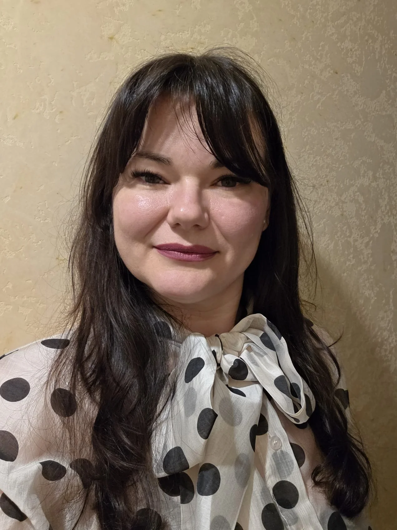 A woman with long dark hair, wearing a polka dot blouse with a bow, smiling at the camera against a textured beige wall.