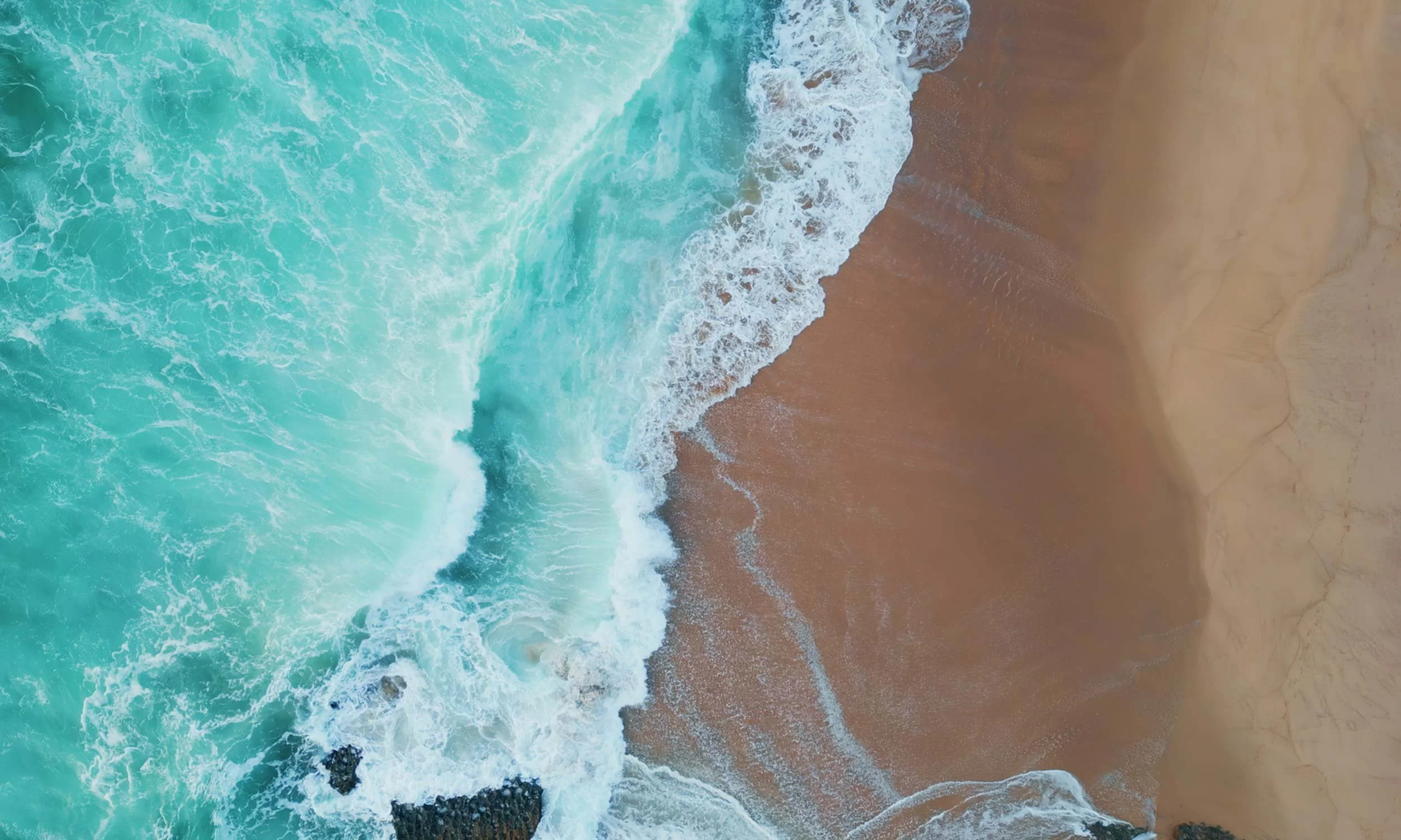 Aerial view of turquoise sea meeting golden sand along a line of white surf, with dark rocks where the foam breaks against the shore.