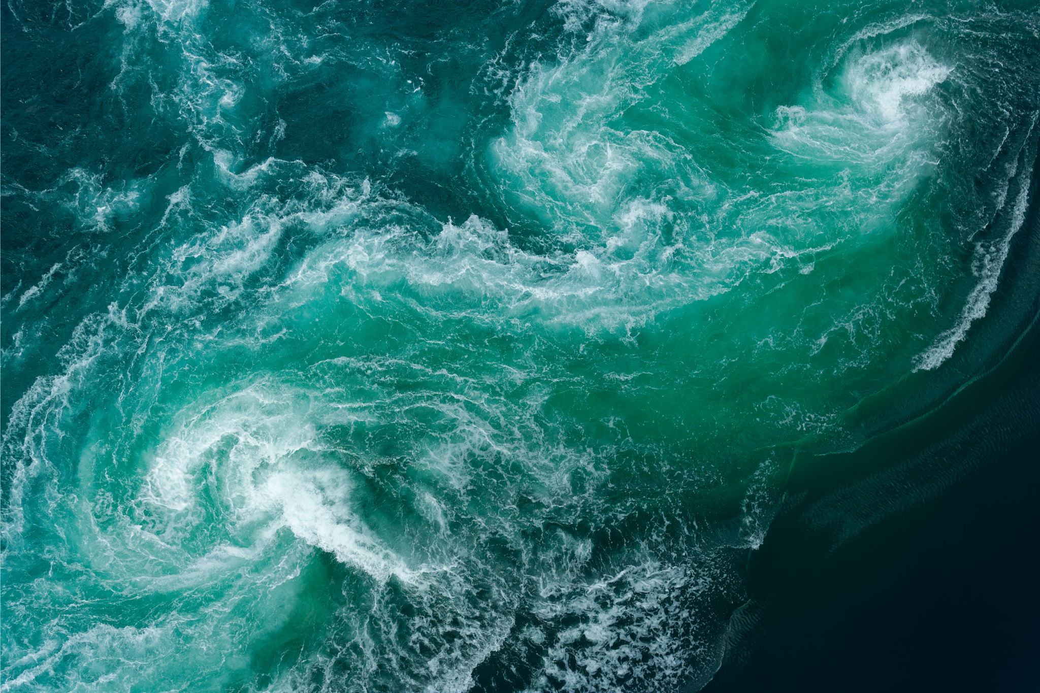Aerial view of swirling teal ocean currents, white foam tracing spirals against dark water.