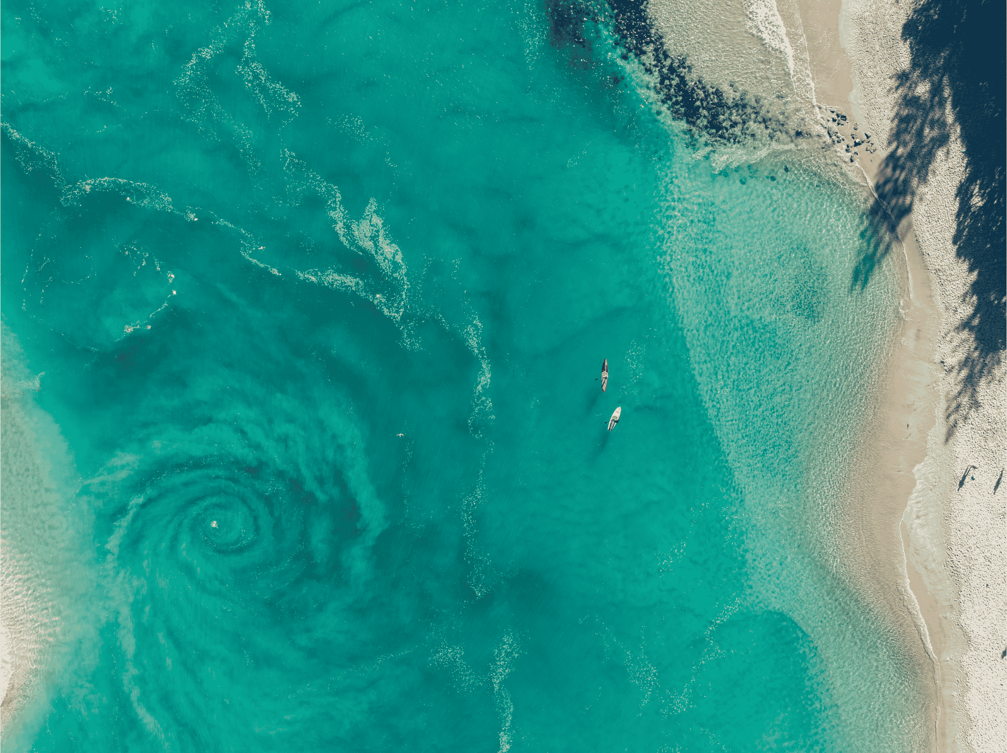 Aerial view of two paddleboarders passing a large spiralling current in clear turquoise water, the spiral visible only from above.