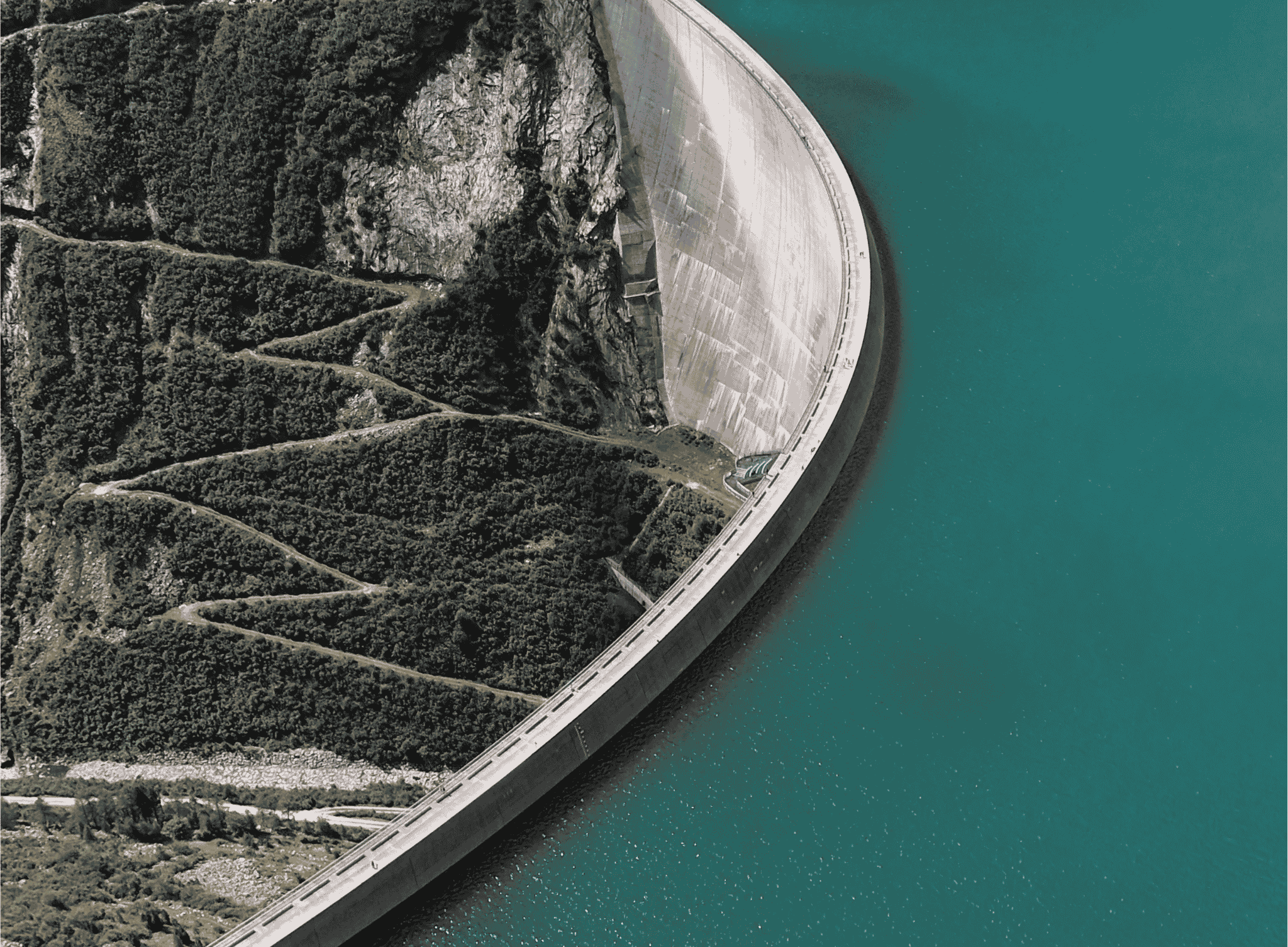 Aerial view of a curved concrete dam holding back a teal reservoir above a green mountain valley, with tiny figures visible on the dam's crest.