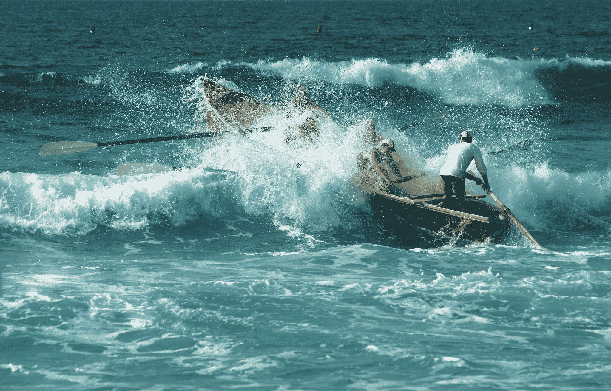A rowing crew in a surf boat driving through a breaking wave, the steerer standing at the stern, the rowers half hidden in spray.