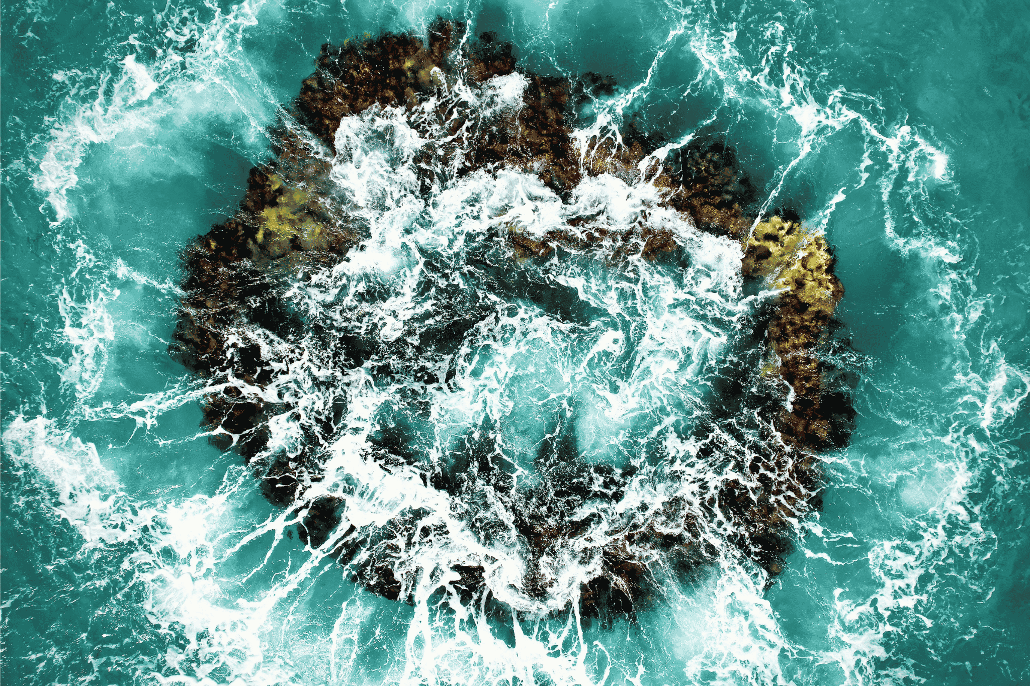 Aerial view of white water surging over a dark rock in turquoise sea, the swell breaking from every side at once.