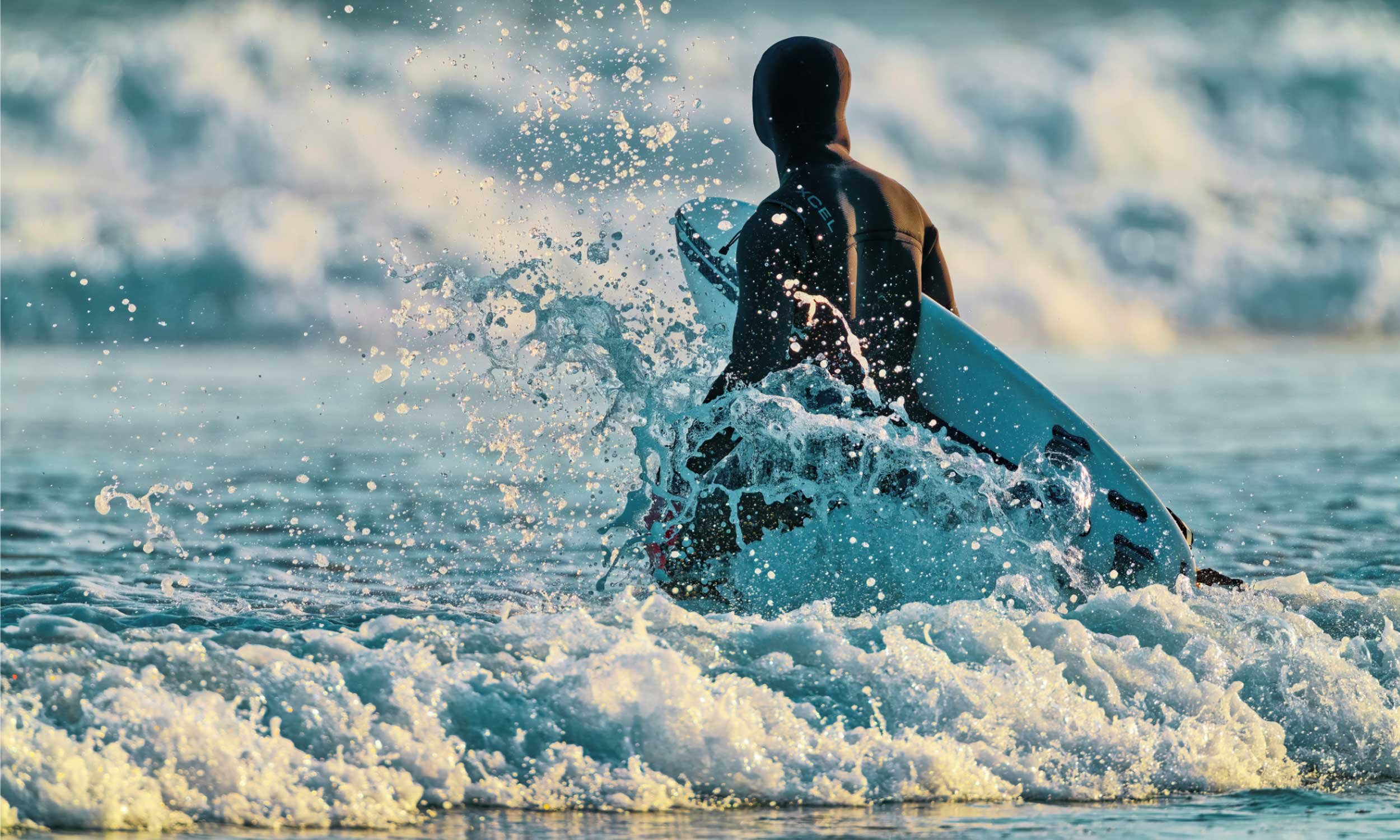 A surfer in a black hooded wetsuit, facing away from camera, sitting on a board pushing through whitewash at sunrise, water spray frozen in the air.