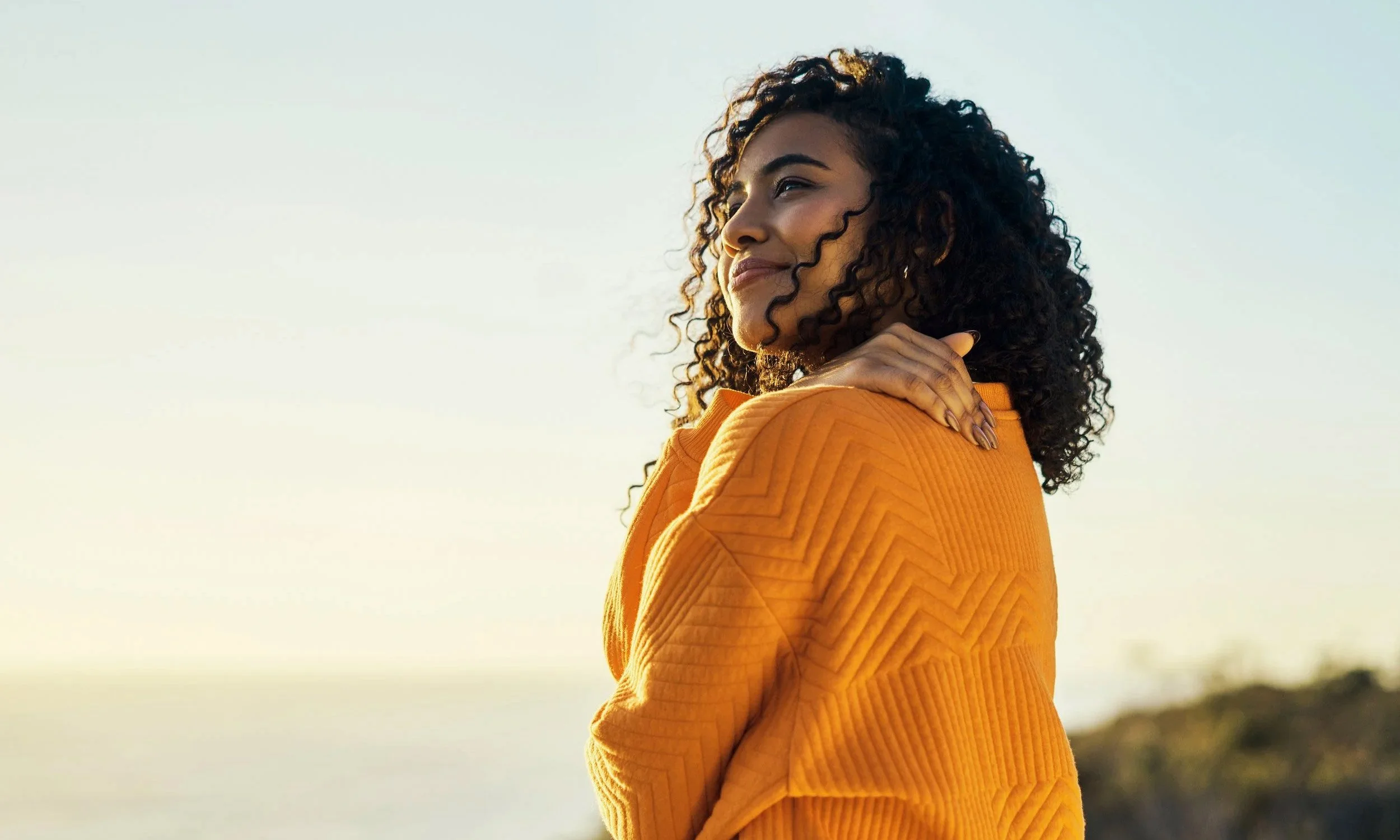 A woman in an orange jumper looks out into a golden sunset, one hand resting on her shoulder.